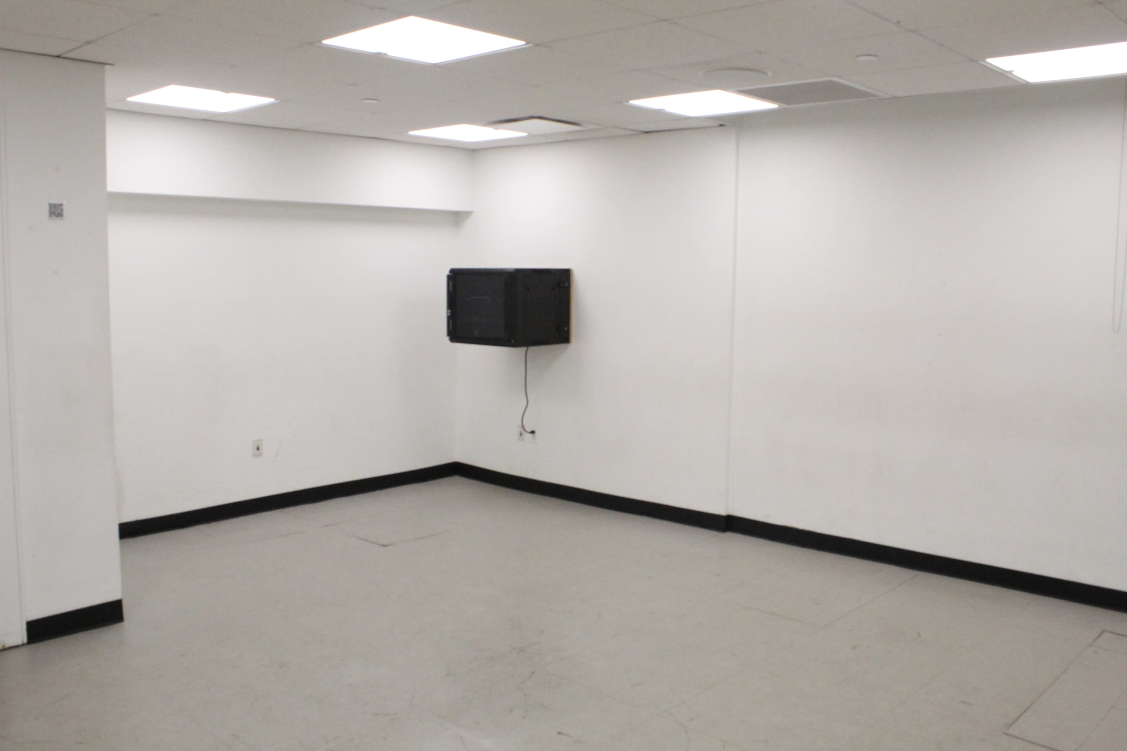 The image shows a sparsely furnished room with white walls and a black display mounted on one wall.