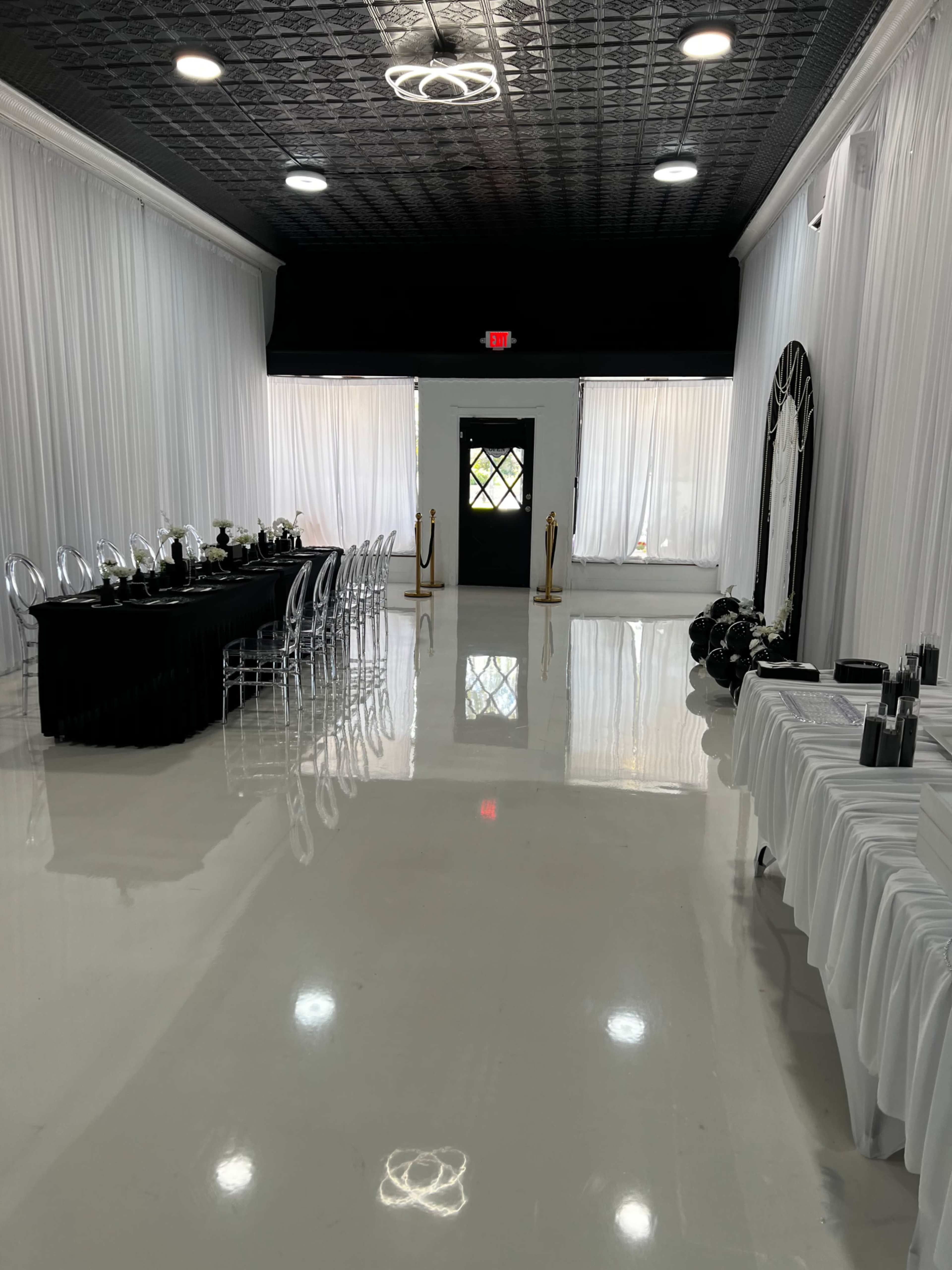 The image shows a spacious event room featuring a long black table set for dining, surrounded by clear plastic chairs, with white curtains and a shiny floor.