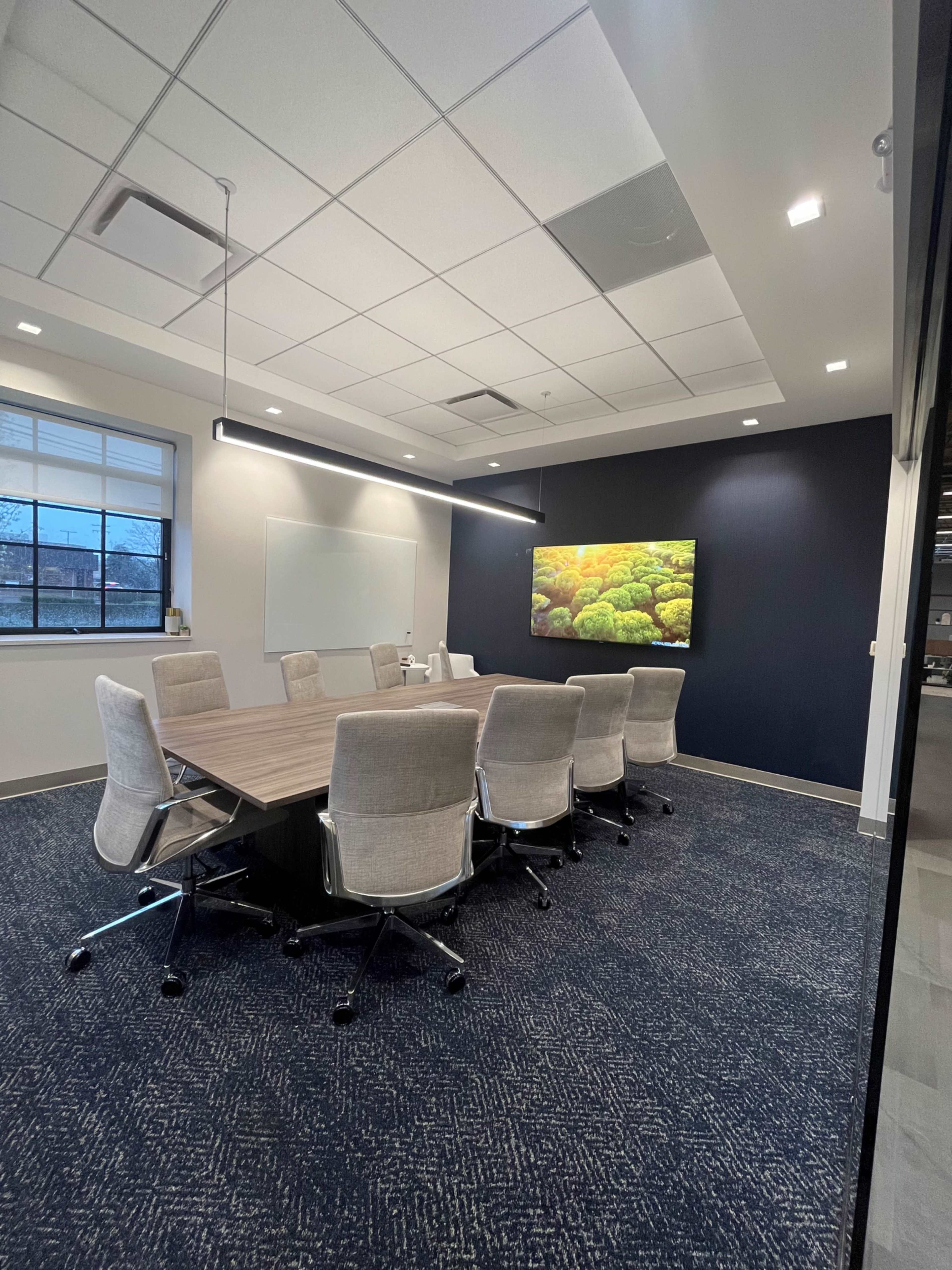 A modern conference room features a large table surrounded by ergonomic chairs, a whiteboard, and a wall-mounted screen displaying a colorful image.