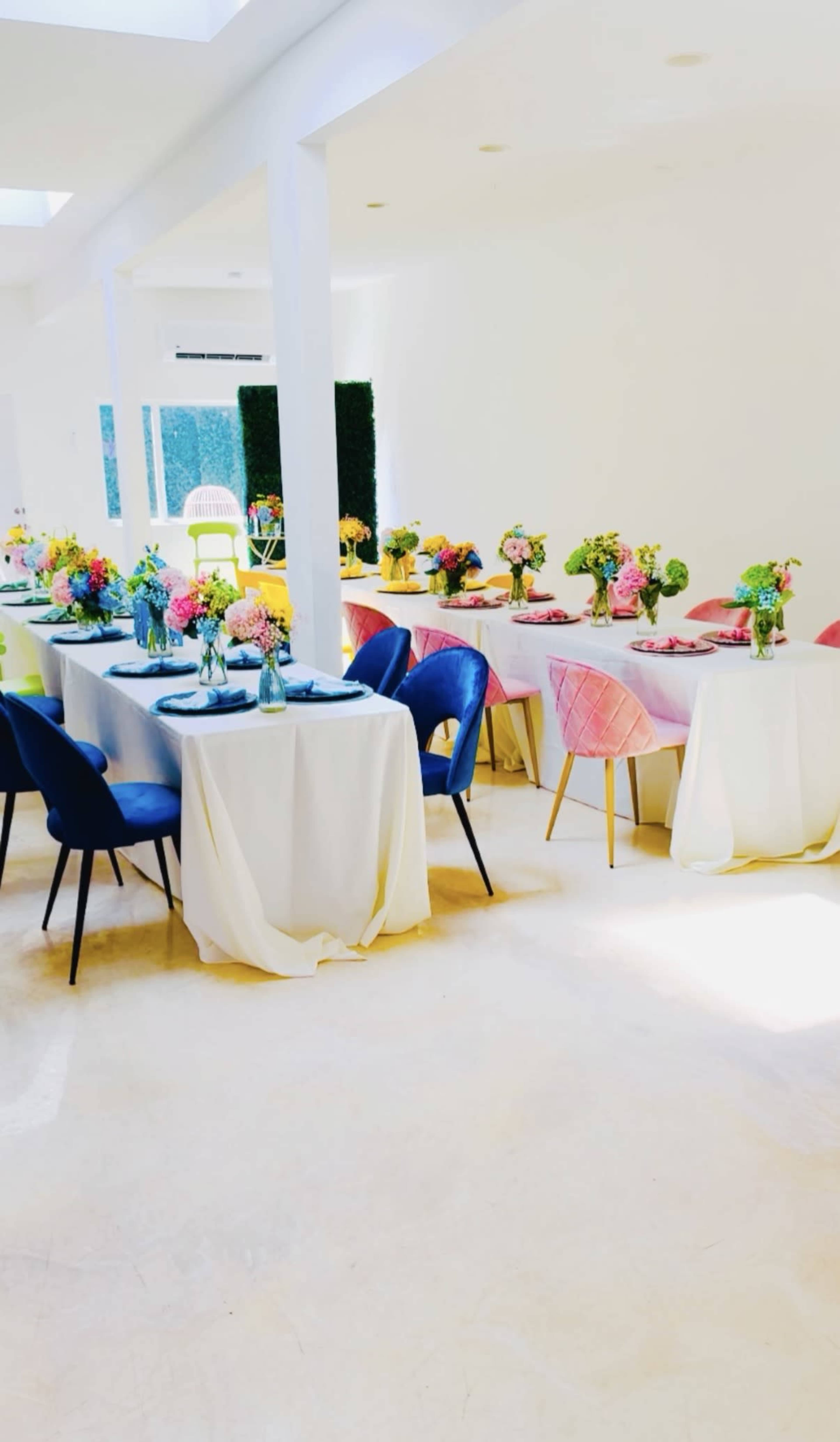 The scene features a brightly lit dining area with multiple tables covered in white tablecloths, decorated with vases of colorful flowers and surrounded by a mix of blue, pink, and yellow chairs.