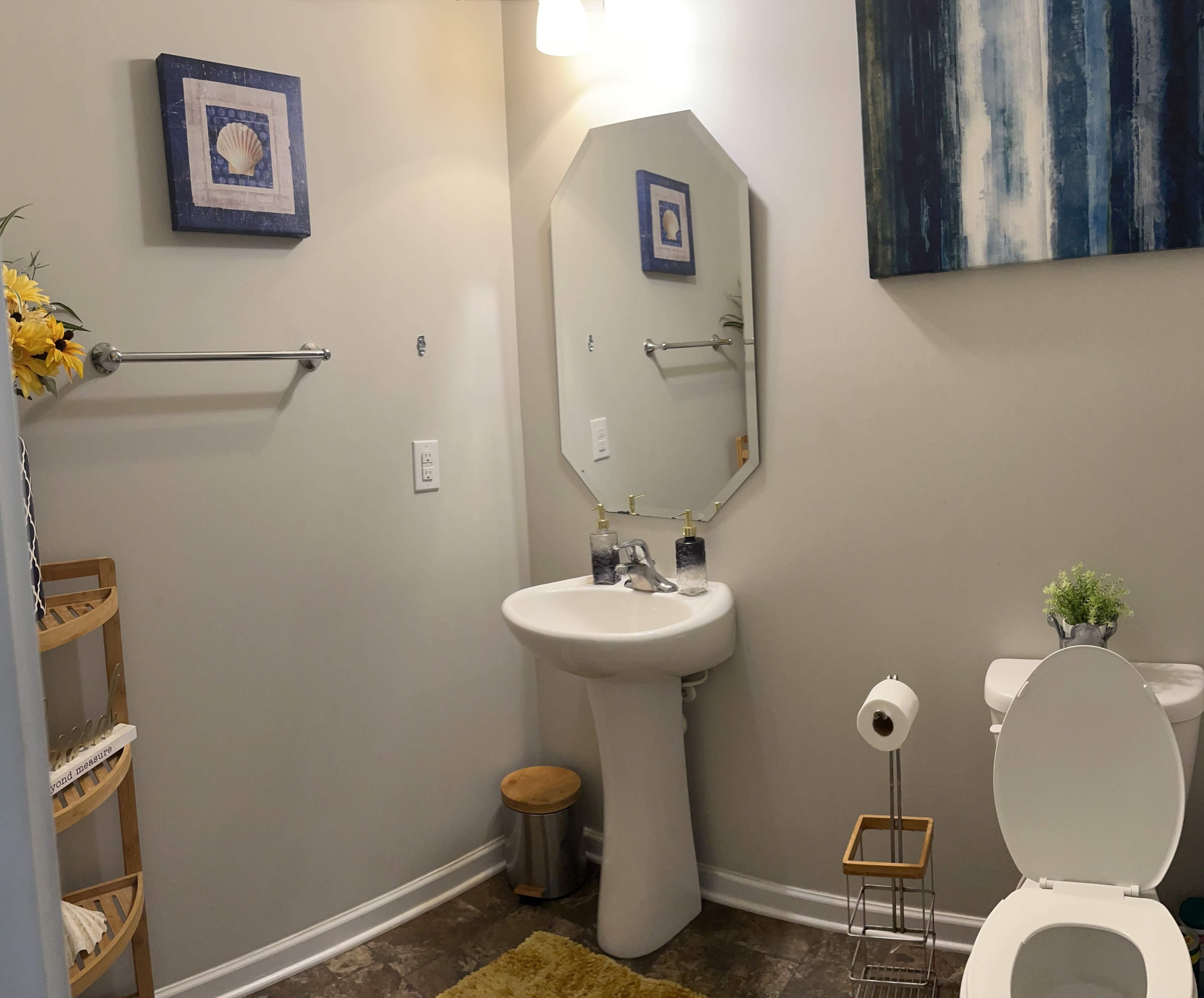 A bathroom featuring a pedestal sink, a mirror, a small wooden stool, and a toilet, with decorative items on the wall and a yellow bath mat on the floor.