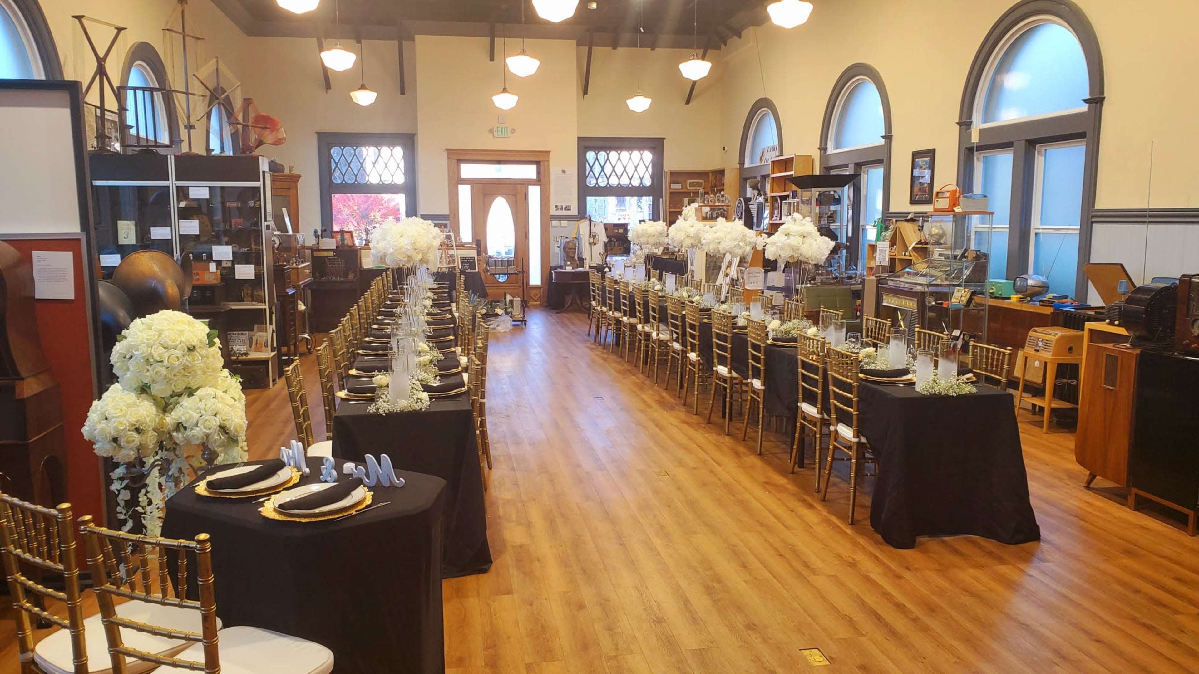 The image shows a spacious indoor venue set up for an event with tables dressed in black cloth and decorated with white floral centerpieces.