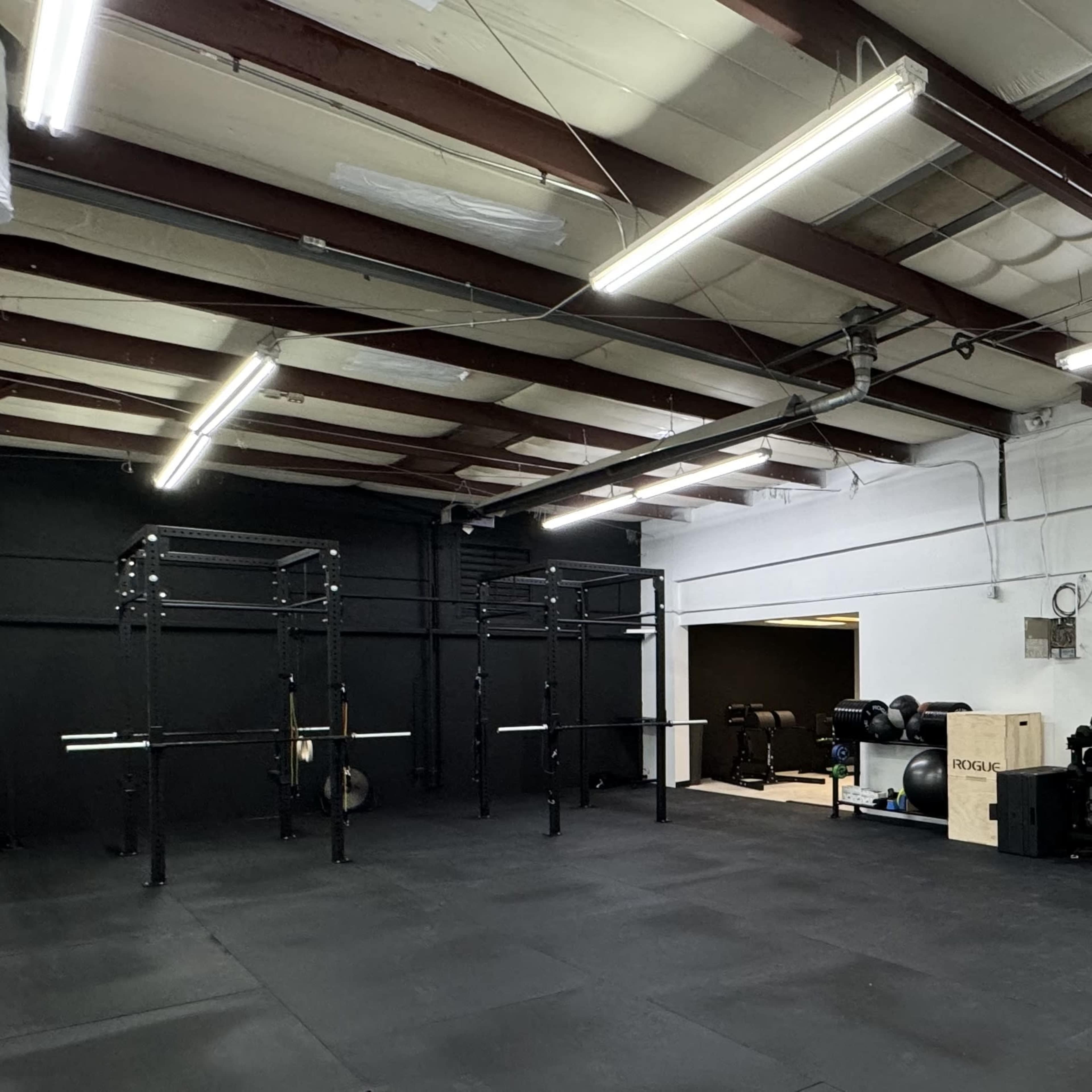 The image shows an empty gym space with a black floor, metal weight racks on one side, and an entrance leading to another room with fitness equipment.