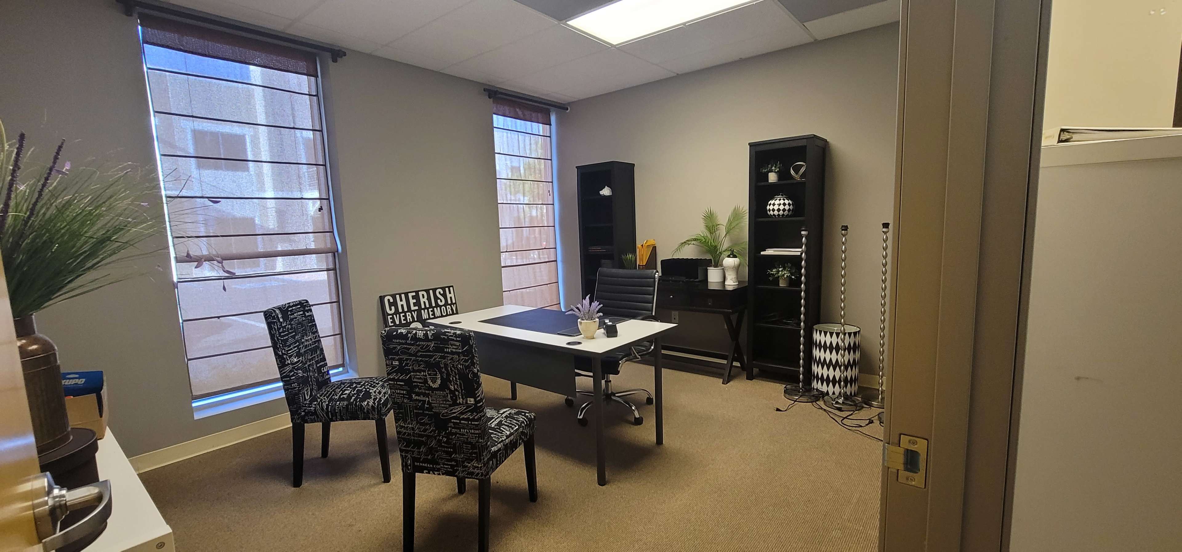 An office space features a desk with a chair, two patterned chairs, a bookshelf, and large windows allowing natural light.