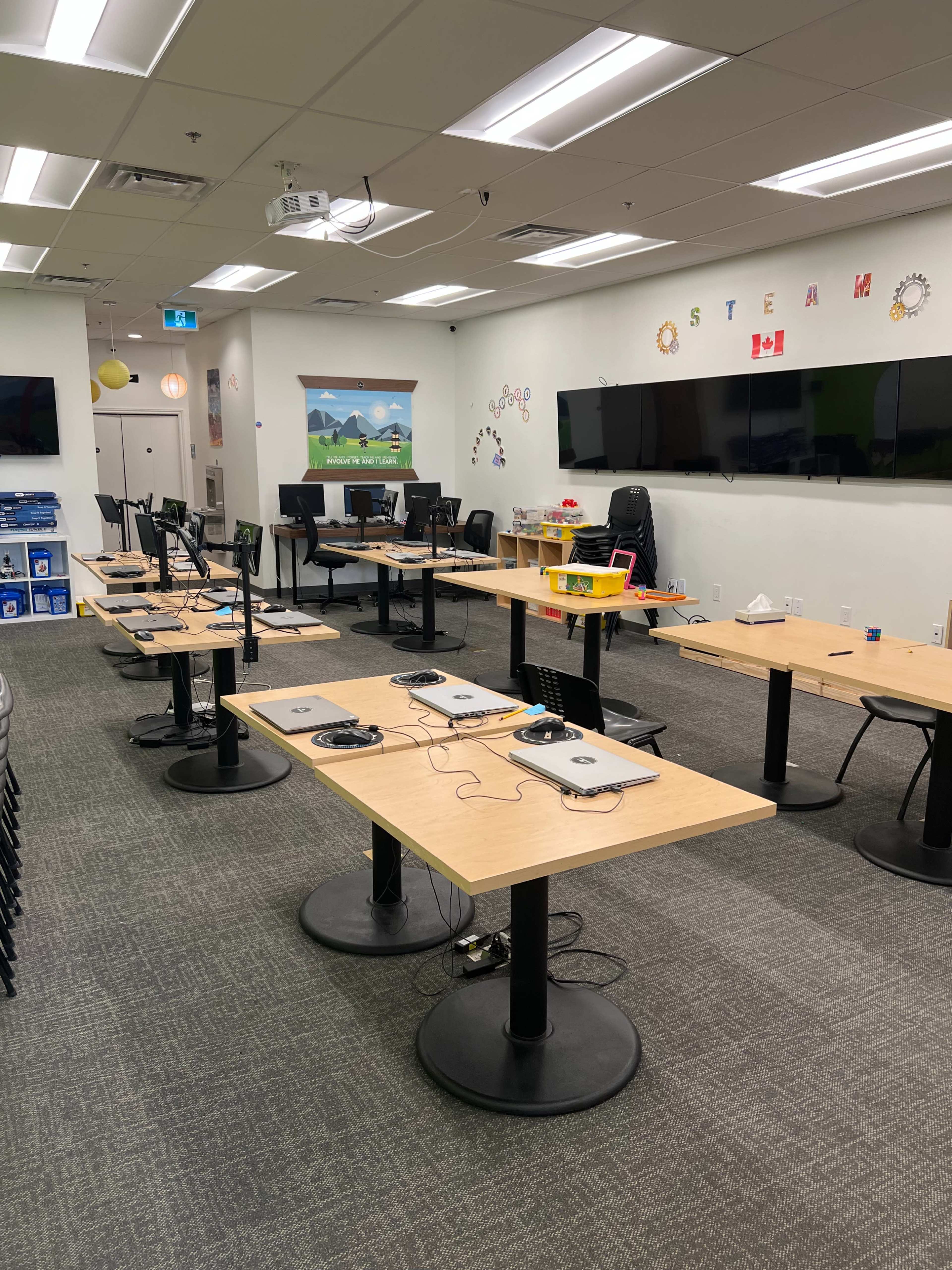 A classroom setup with several tables arranged in a grid, each equipped with laptops, in a brightly lit space featuring educational decorations on the walls.