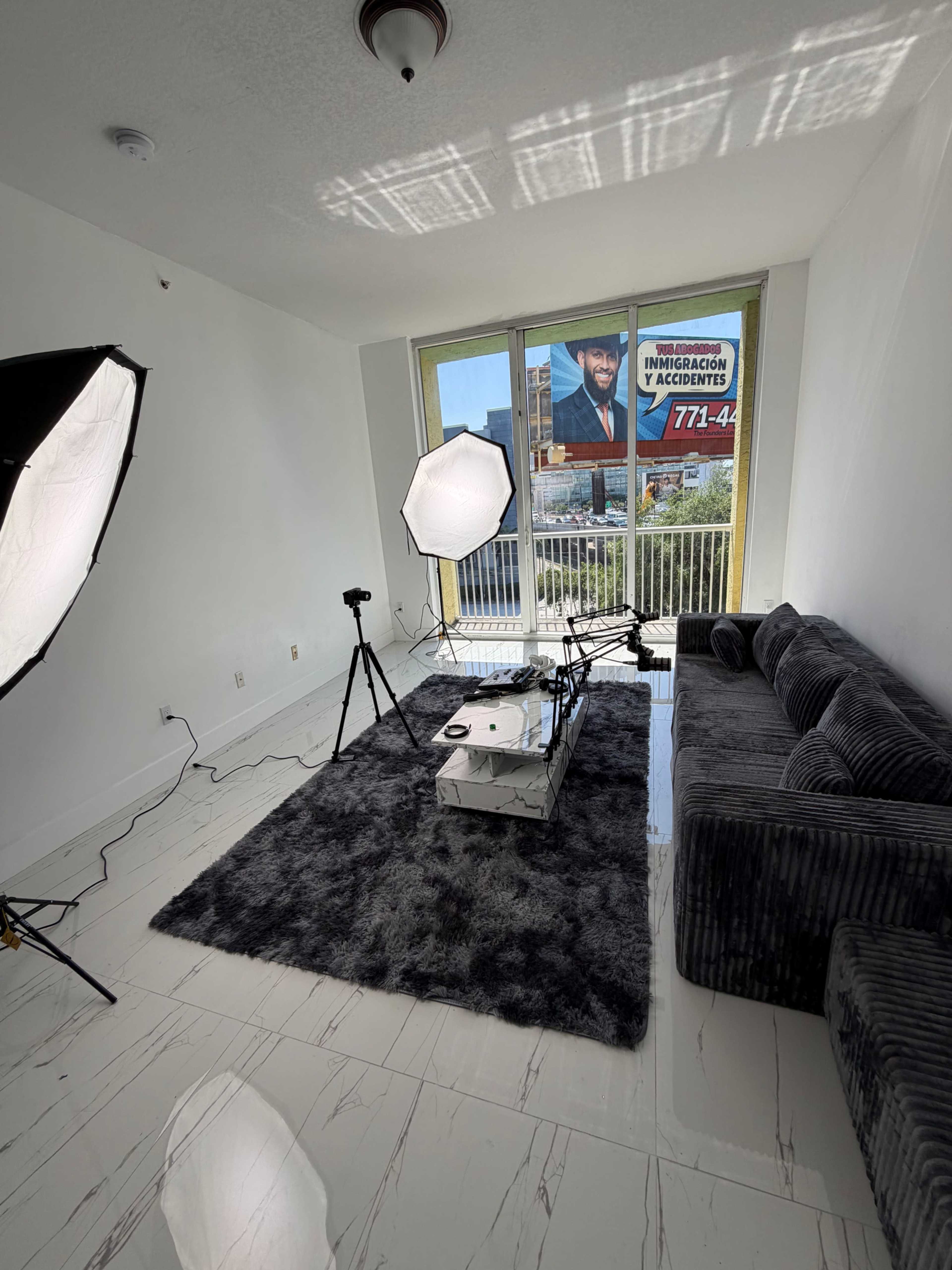 The image shows a modern living room setup with a gray sofa, a coffee table, and photography equipment including lights and a camera on tripods, with a large advertisement visible through the window.