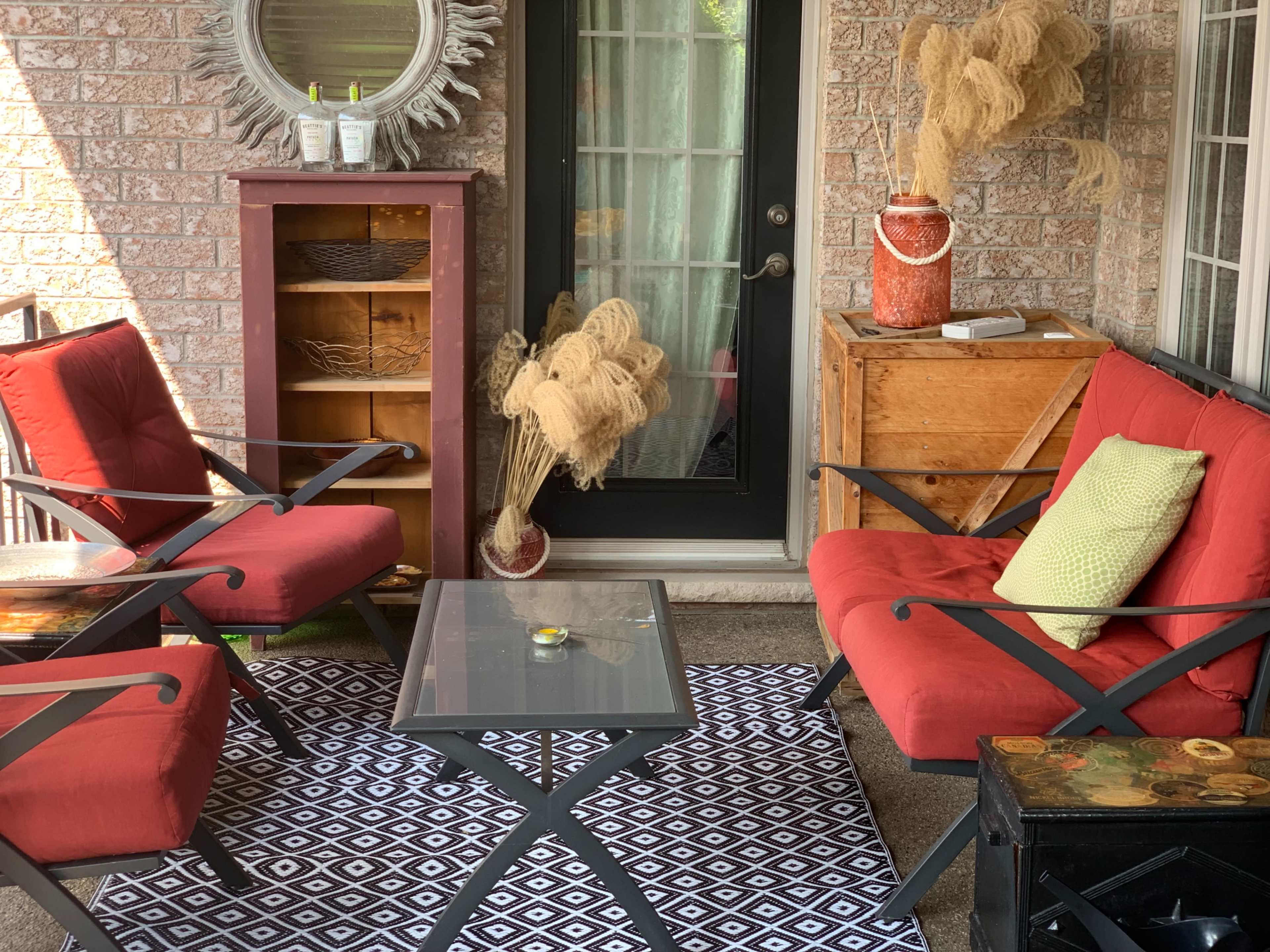 A cozy porch area featuring two red chairs, a glass table, a patterned rug, and decorative elements like a mirror and potted plants.