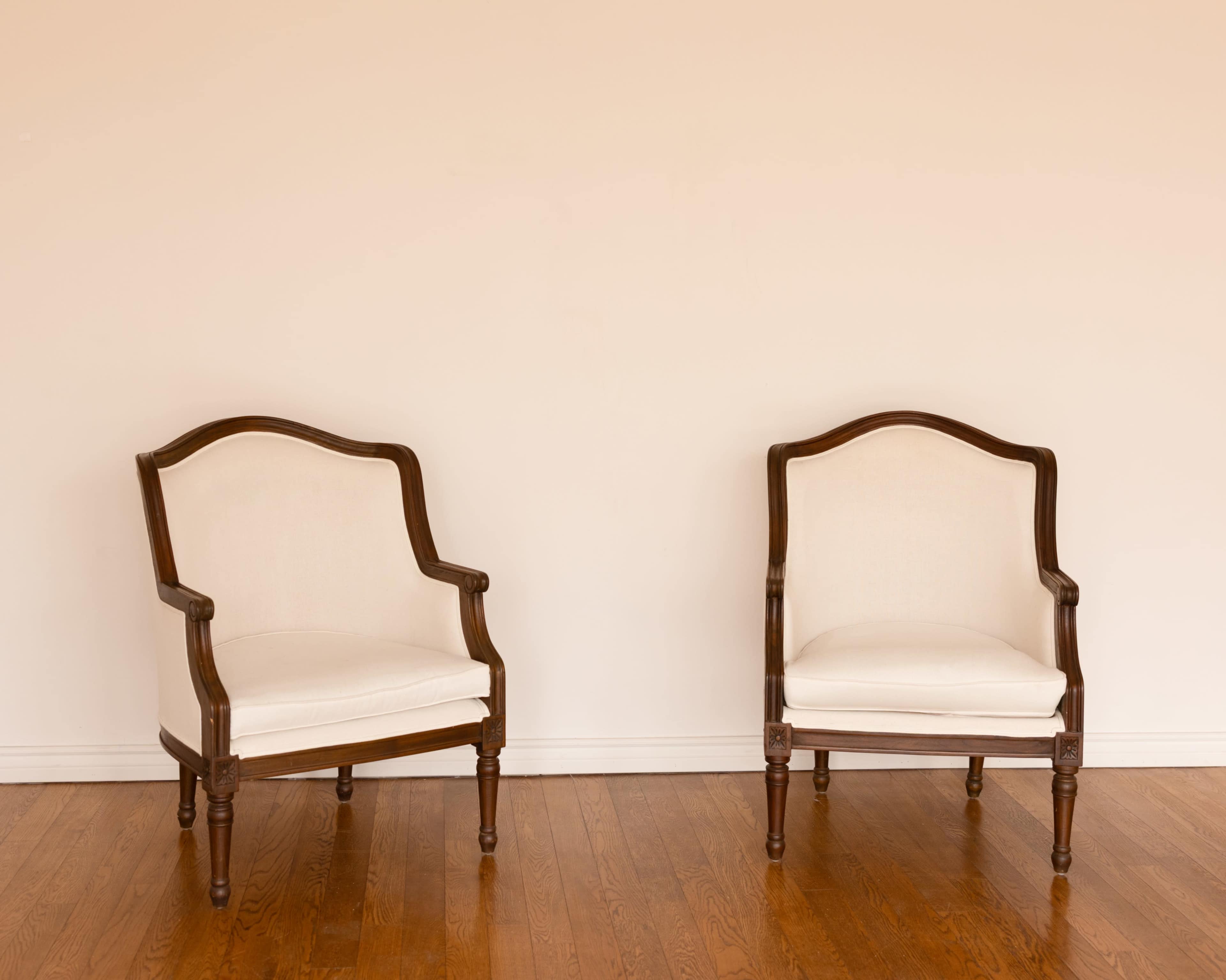 Two wooden armchairs with light-colored fabric upholstery are placed facing each other on a wooden floor against a plain wall.