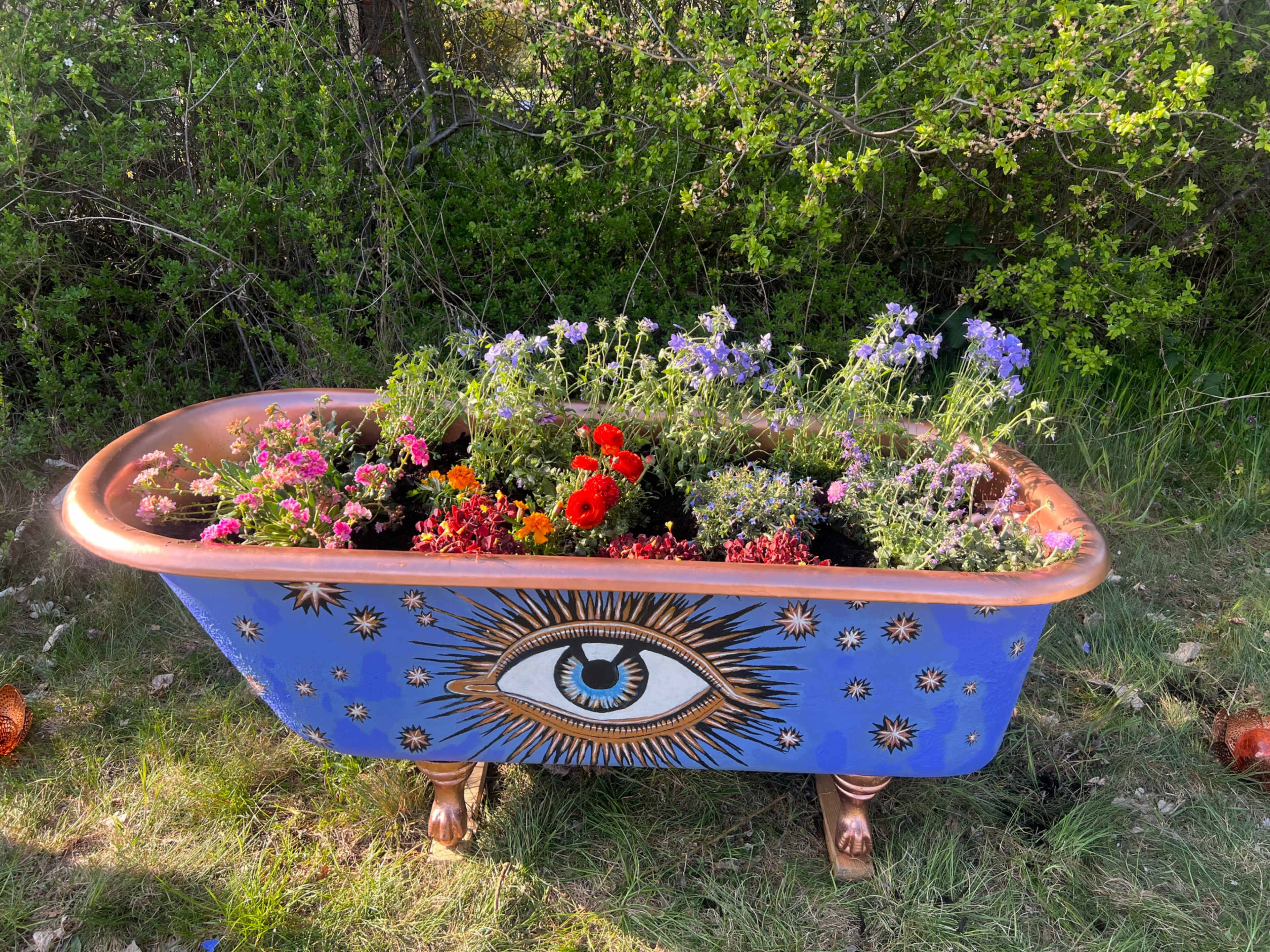 A brightly painted vintage bathtub filled with colorful flowers is set against a backdrop of greenery.