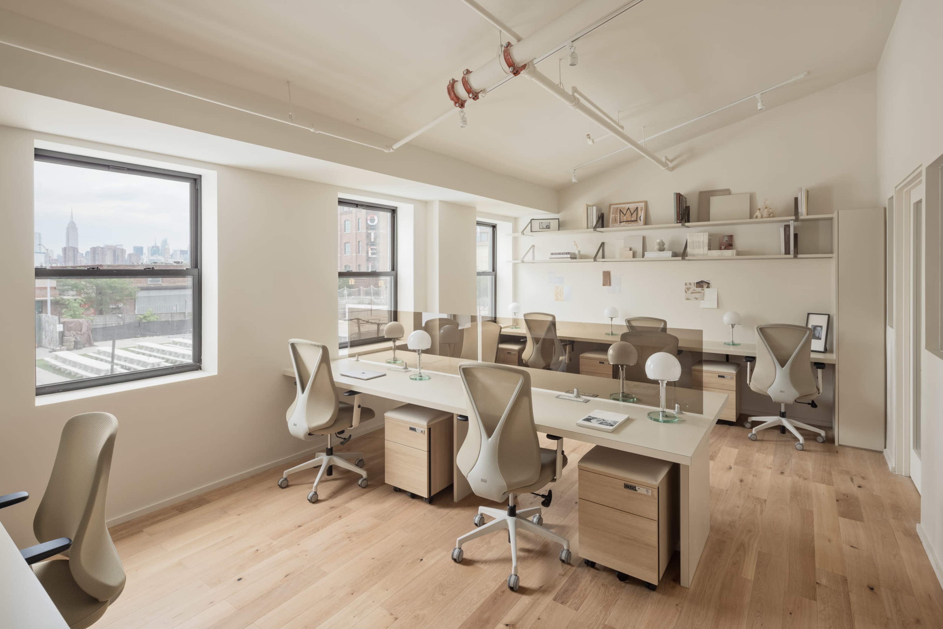 The image shows a modern office space with several workstations, large windows, and wooden flooring, featuring minimalist furniture and a view of a city skyline.