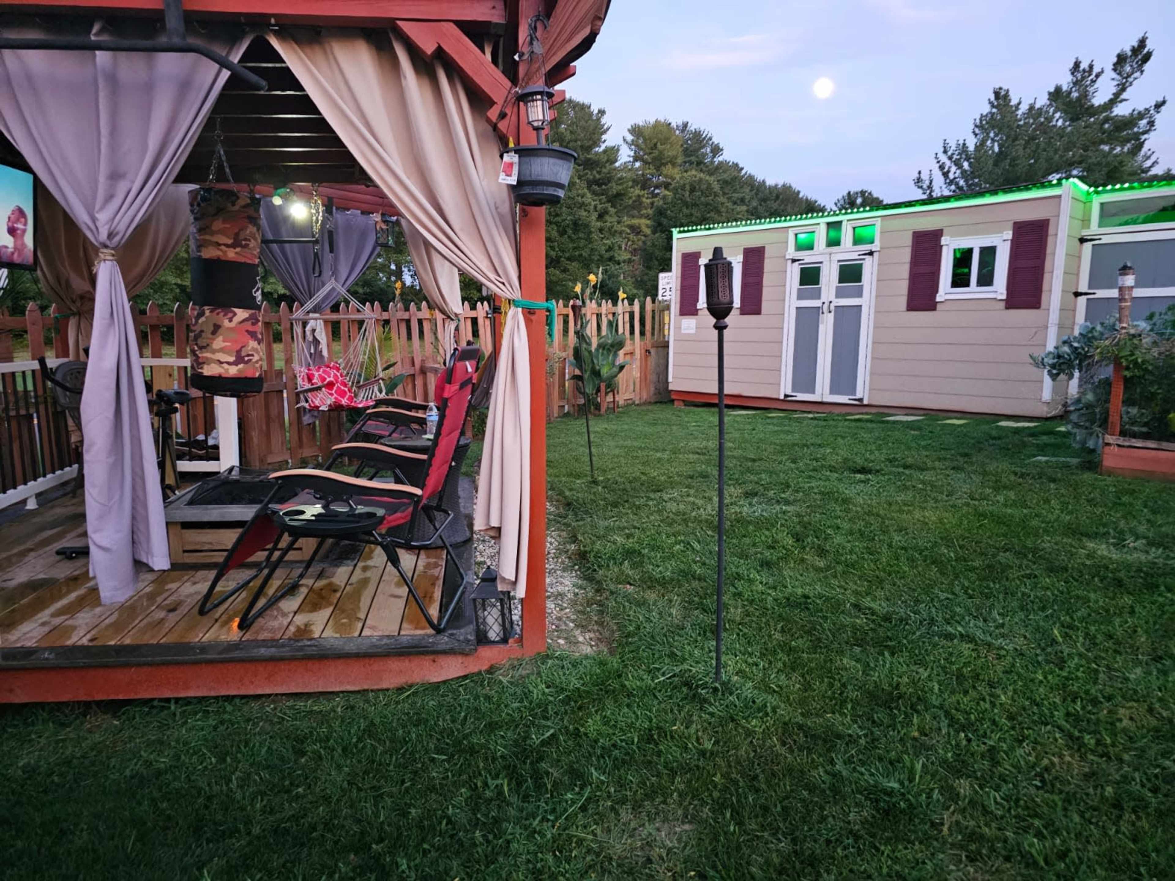 A wooden outdoor patio with chairs and draped curtains is adjacent to a small shed outlined with green lights in a grassy yard.