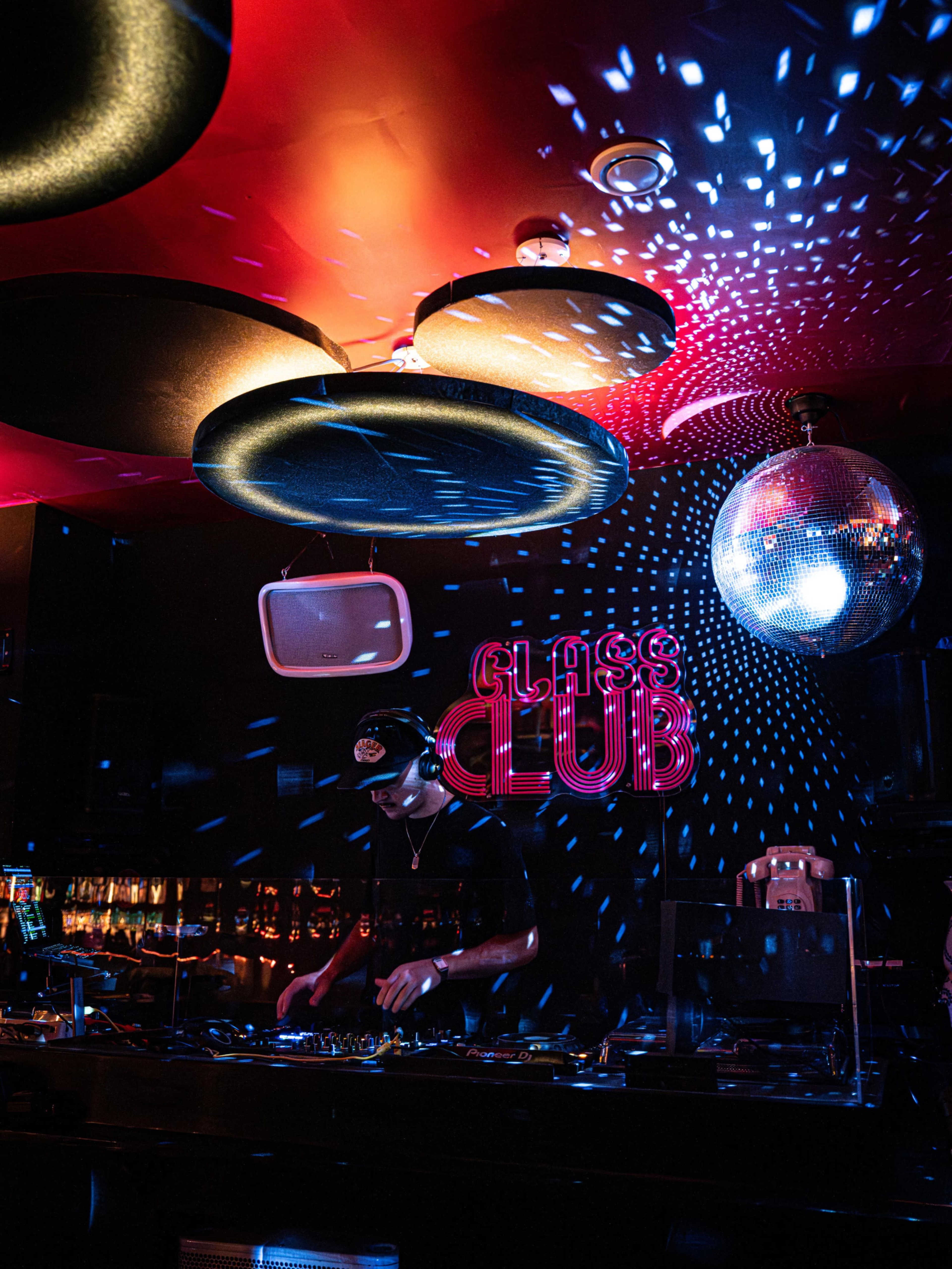 A DJ stands at a booth in a nightclub filled with colorful lighting and a disco ball reflecting patterns on the walls.