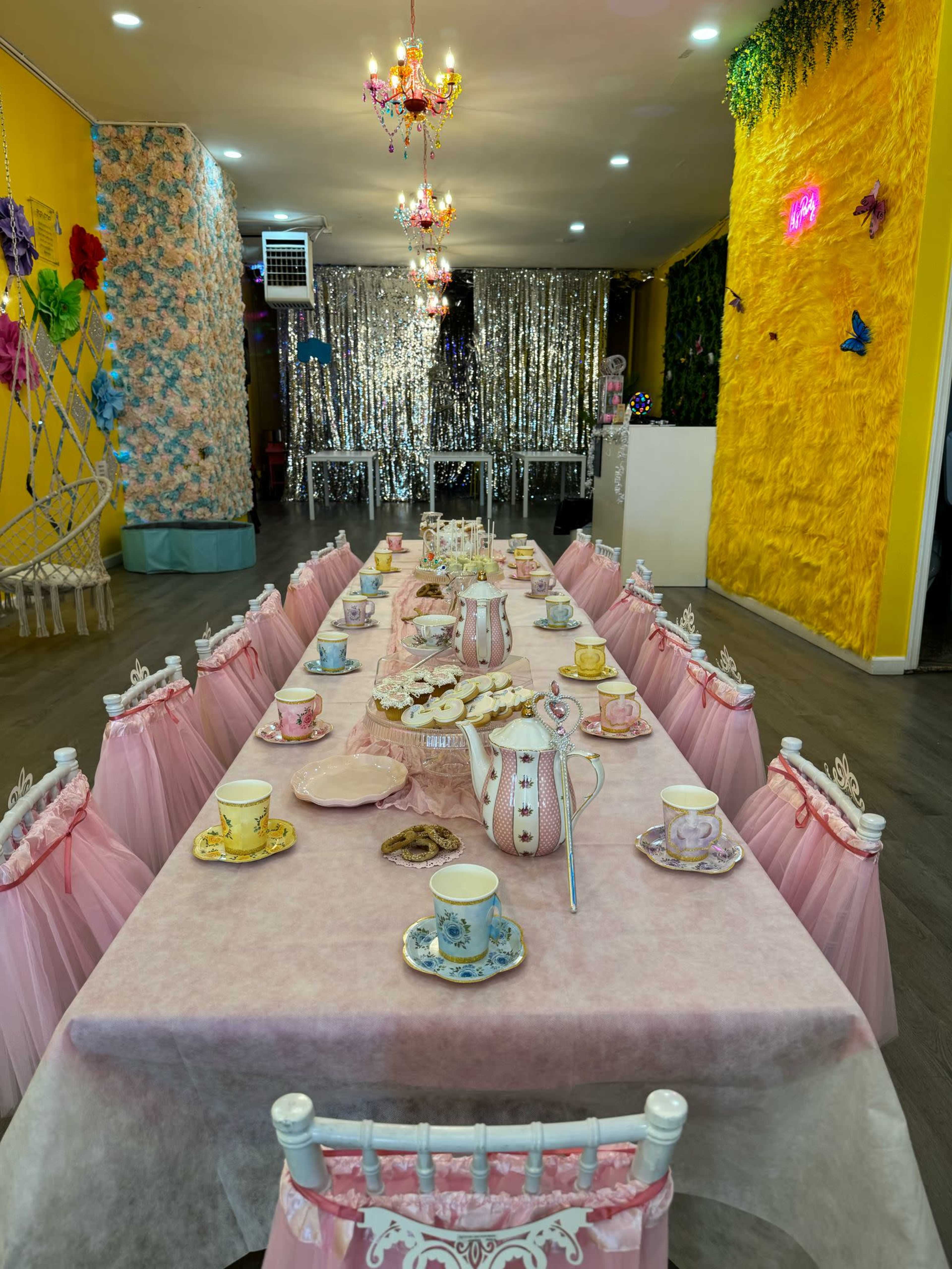 The image shows a decorated dining area with a long table set for a tea party, featuring pink tablecloths, ornate chairs, and various cups and dishes arranged neatly on the table.