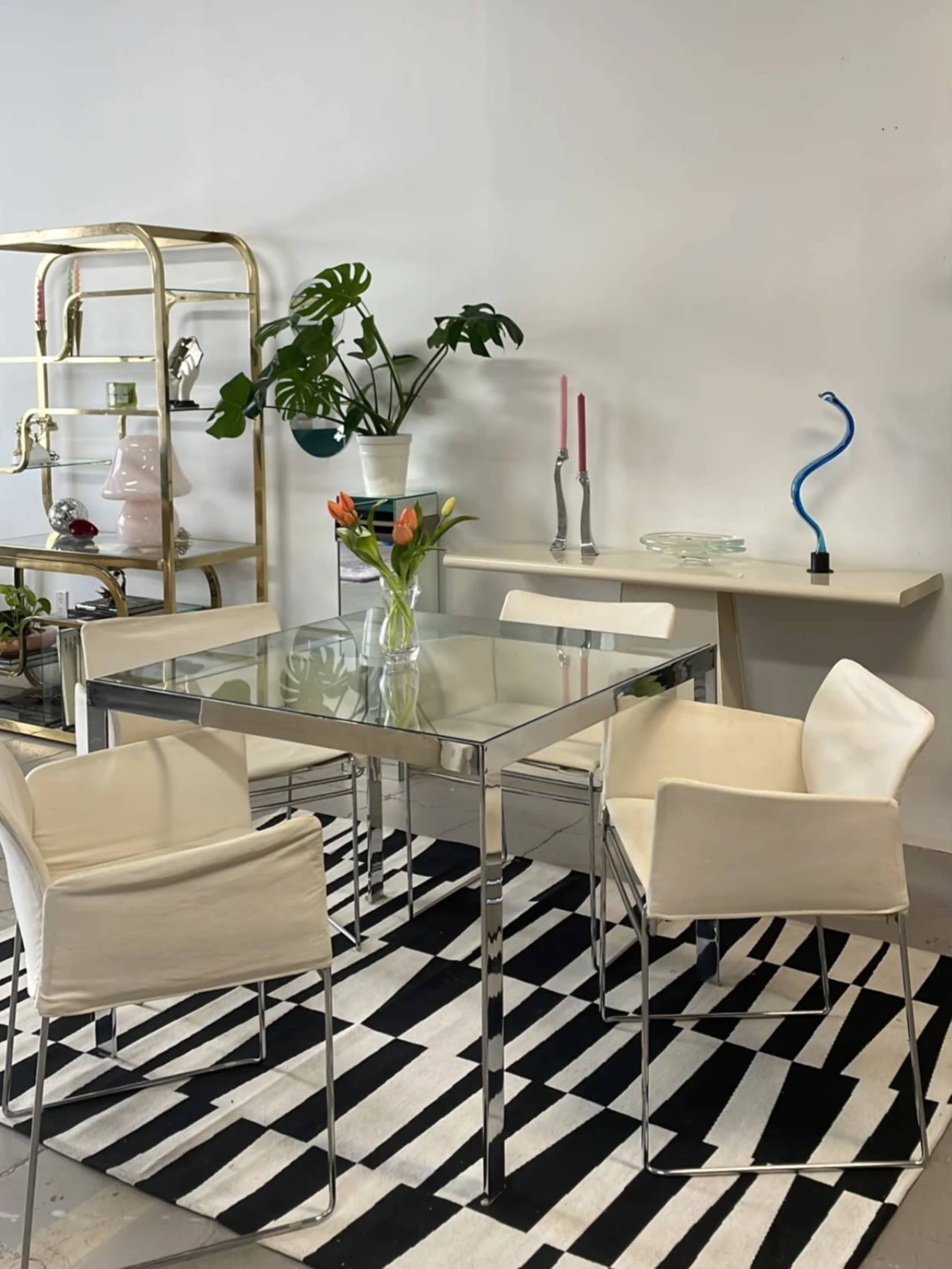 A glass-top table with four white chairs is set on a black and white striped rug, accompanied by a plant, candles, and decorative items in the background.