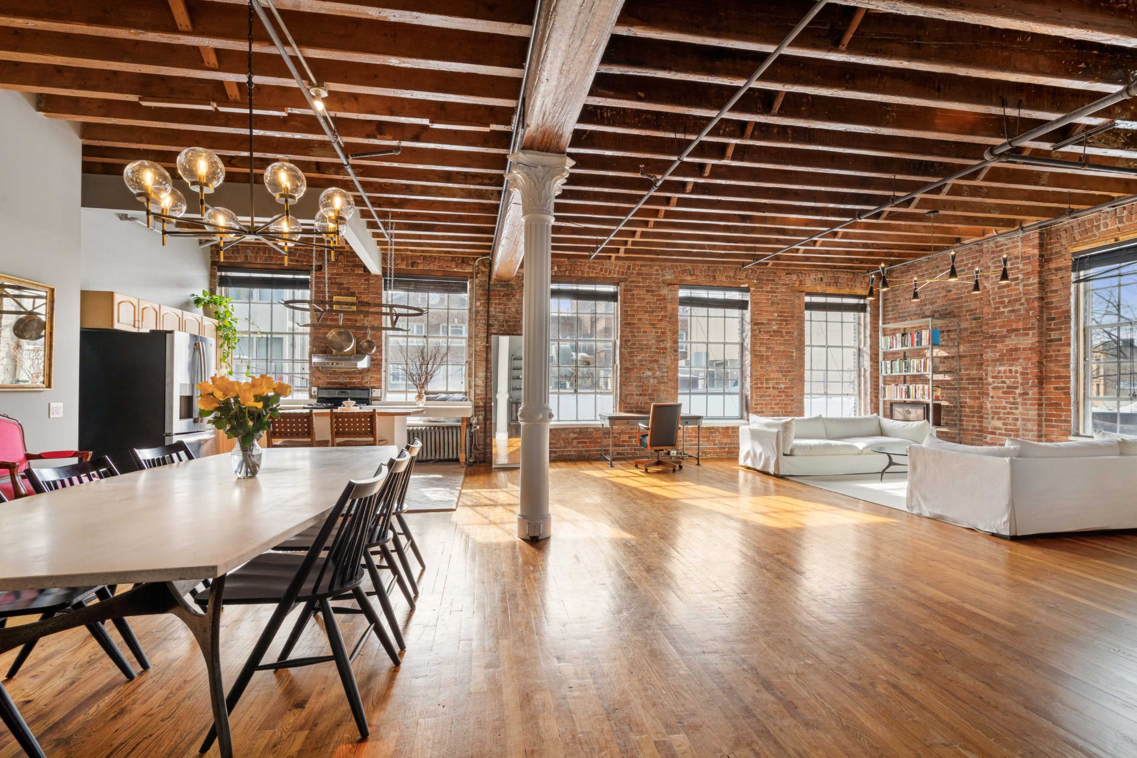 The image shows a spacious loft with exposed brick walls, wooden beams, and large windows, featuring a dining area with a long table and a living area with a sofa.