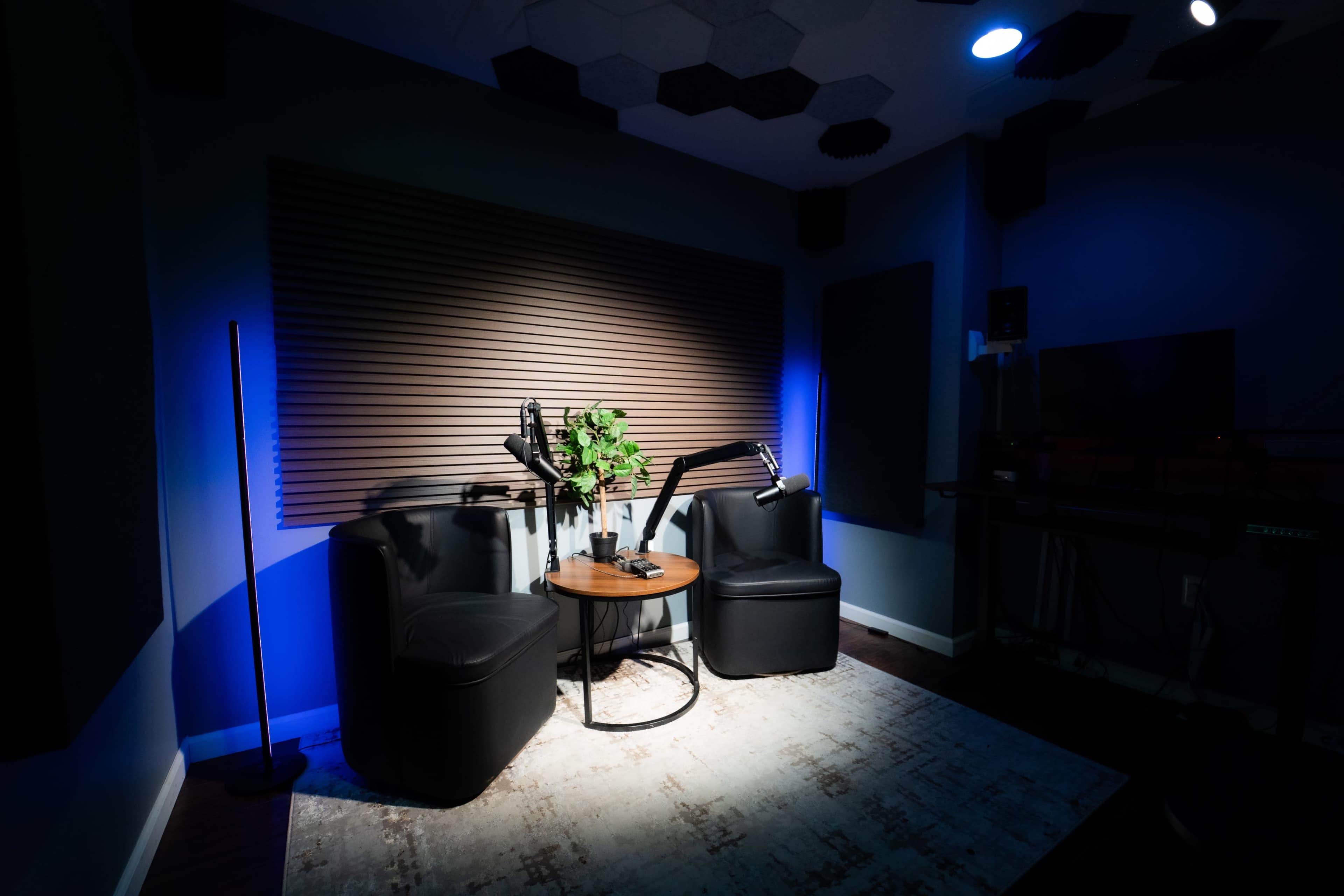 The image shows a small recording studio with two black chairs, a round table, and a potted plant, illuminated by soft lighting.
