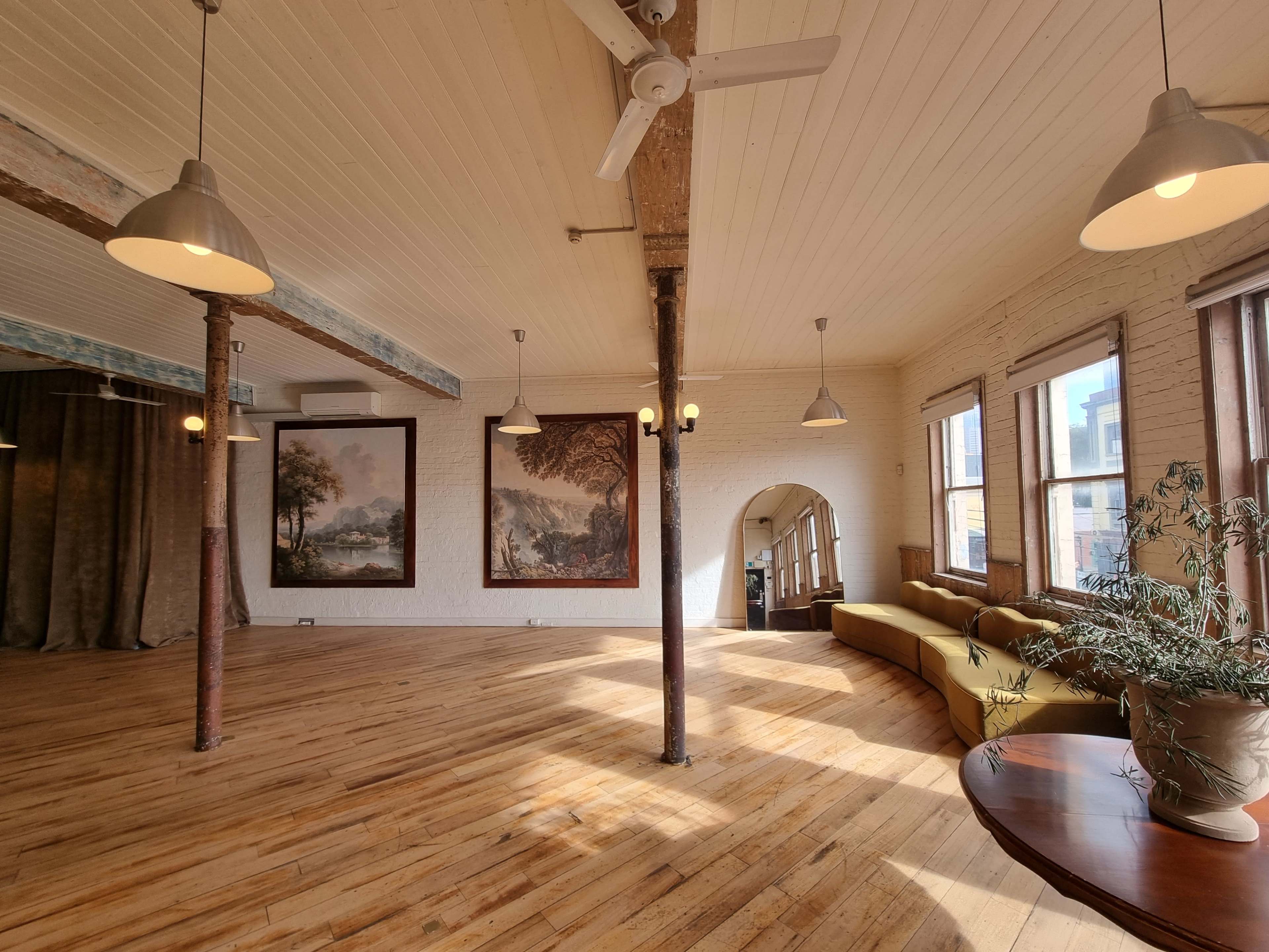 The image shows a spacious, well-lit room with wooden floors, exposed beams, and large paintings on the walls, featuring a sofa and a round table.