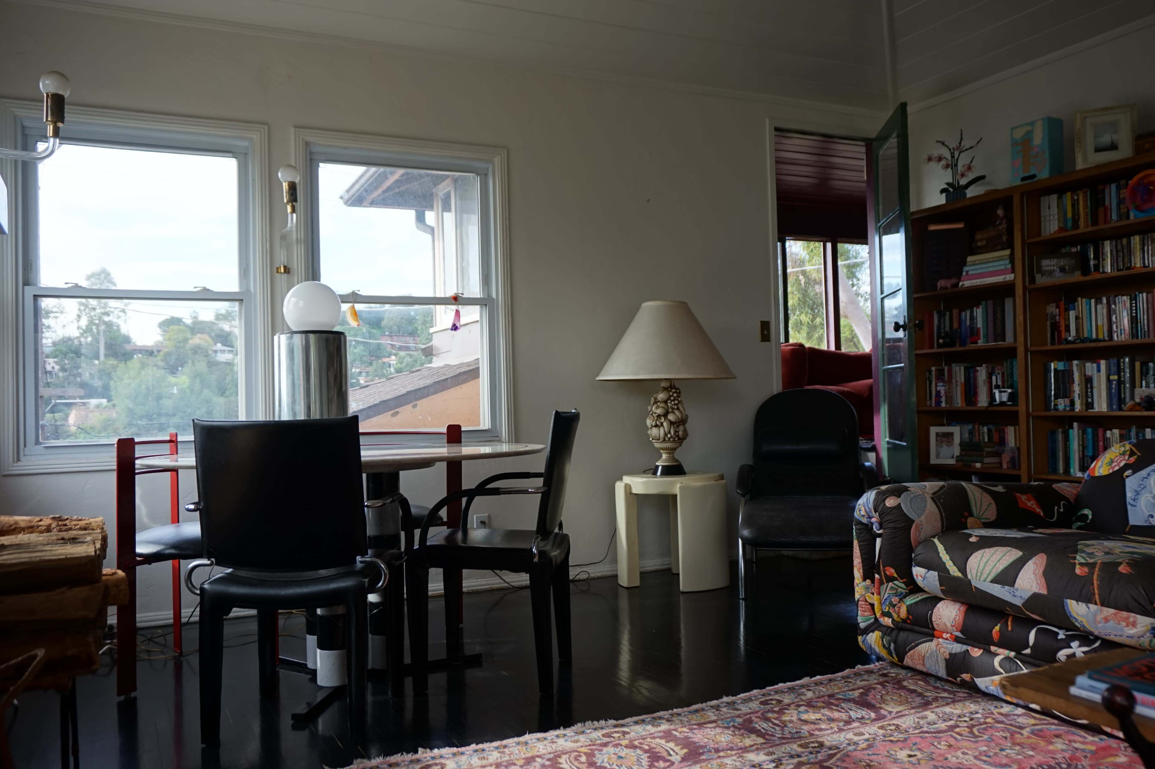The image shows a living space with a dining table and chairs, a bookshelf, a lamp, and a patterned couch, illuminated by natural light coming through large windows.