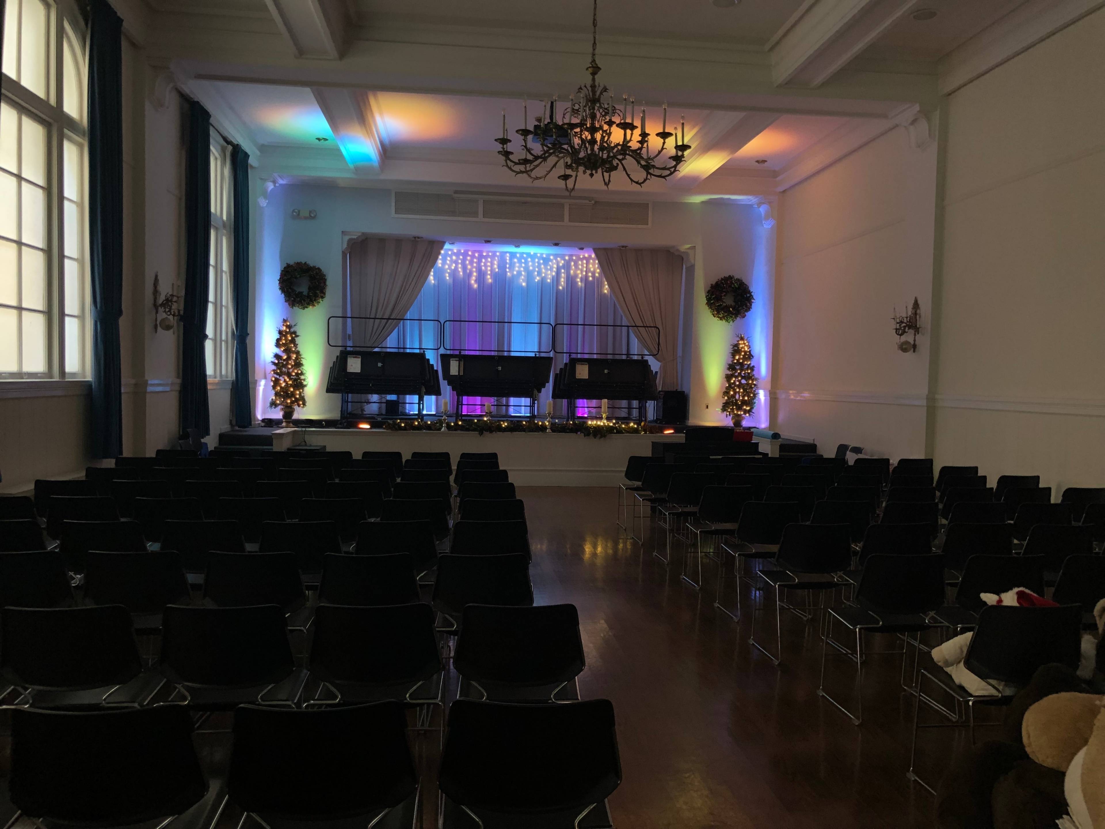 A stage is set with microphones and colorful lighting in a large event hall, surrounded by rows of black chairs and decorated with Christmas trees.