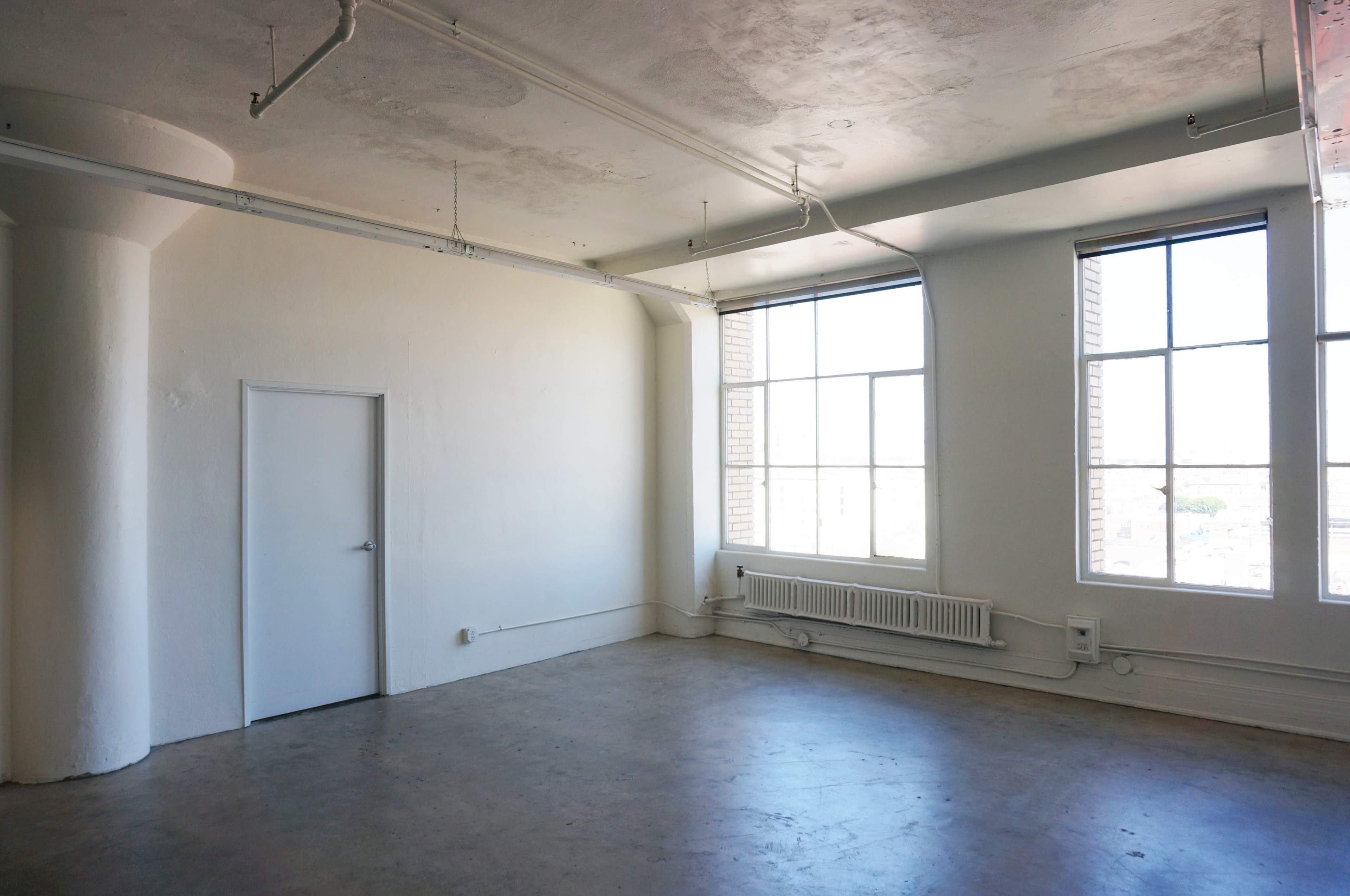The image shows an empty room with large windows, a concrete floor, and a single door.