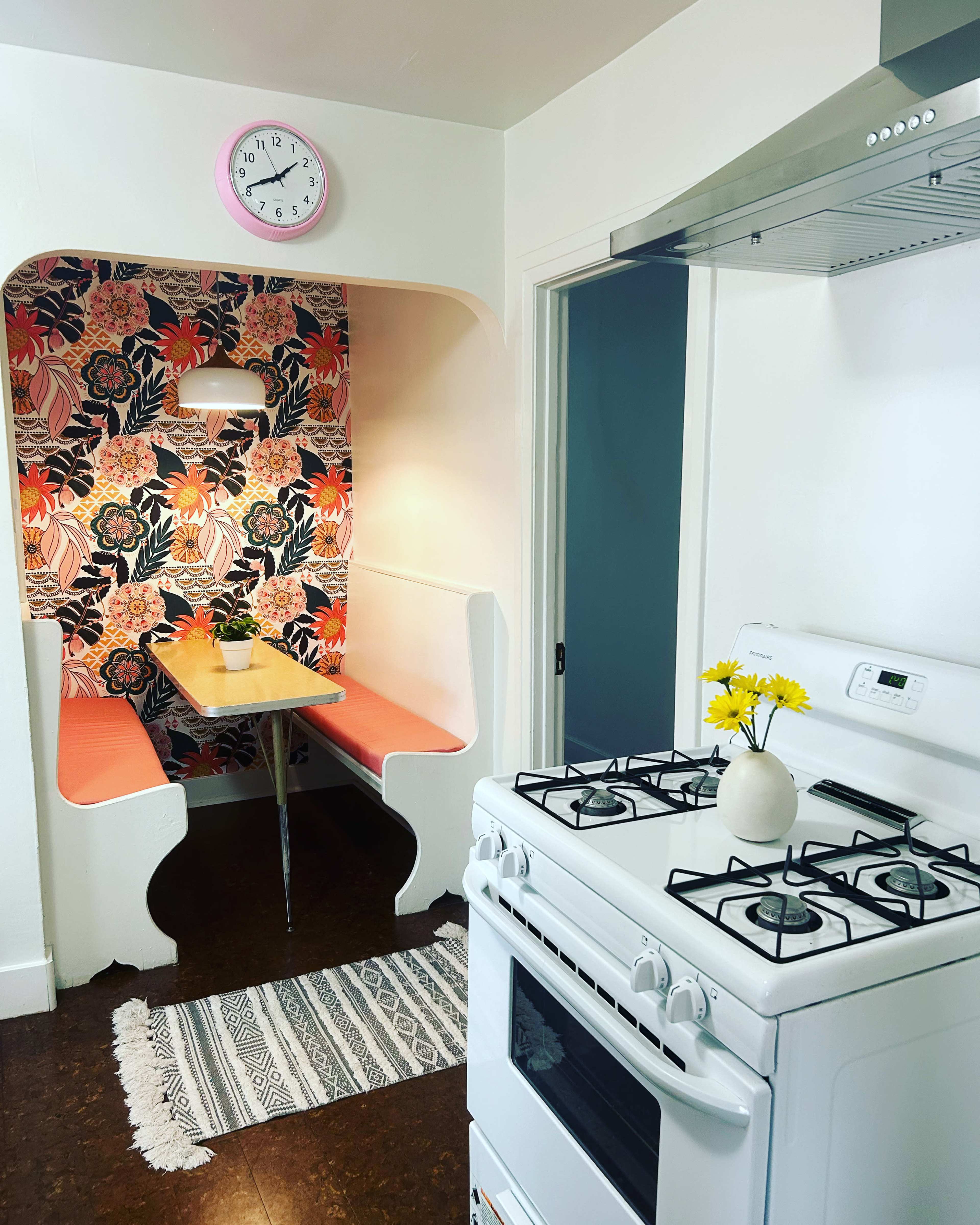 The image shows a kitchen area with a white stove, a decorative floral wallpaper behind a small dining nook, and a table with a vase of flowers.