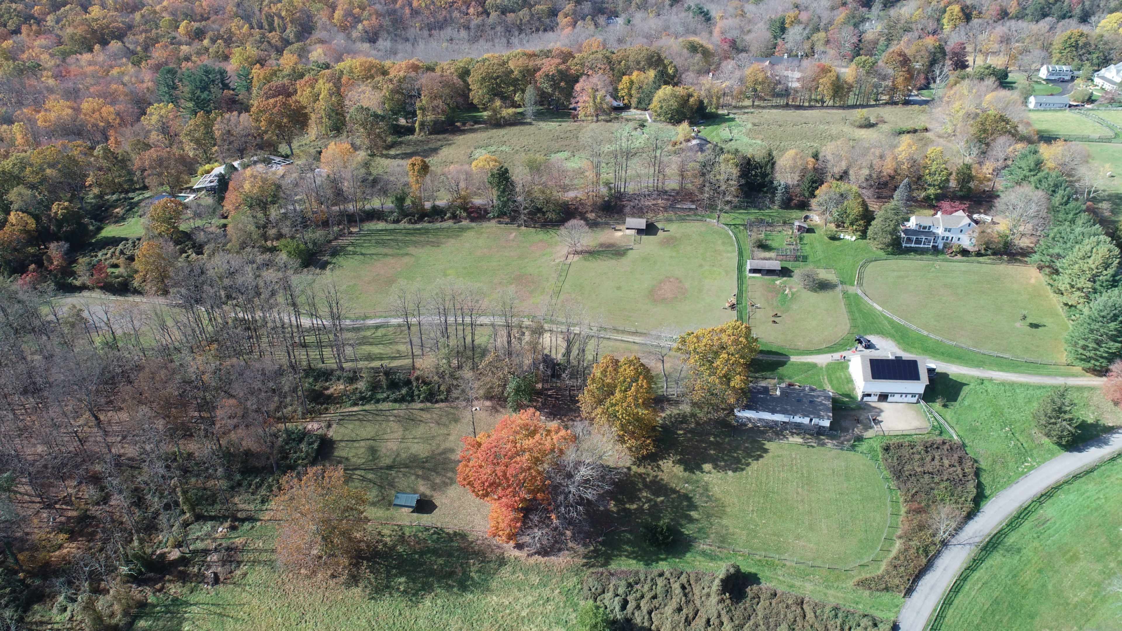 The image shows an aerial view of a rural landscape with autumn foliage, featuring fields, sparse trees, a few structures, and winding roads.