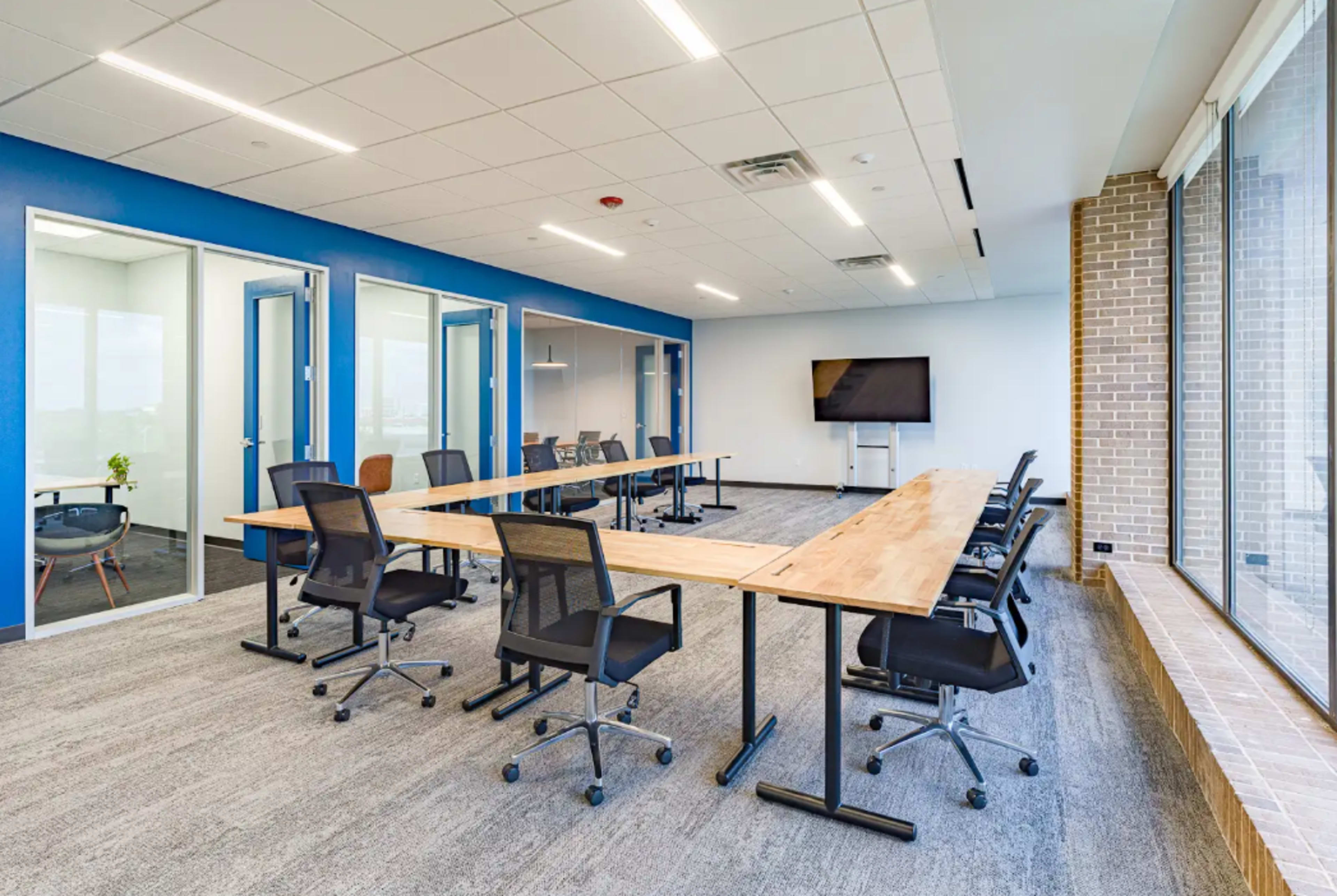 The image shows a modern conference room with a long wooden table surrounded by black office chairs and large windows.
