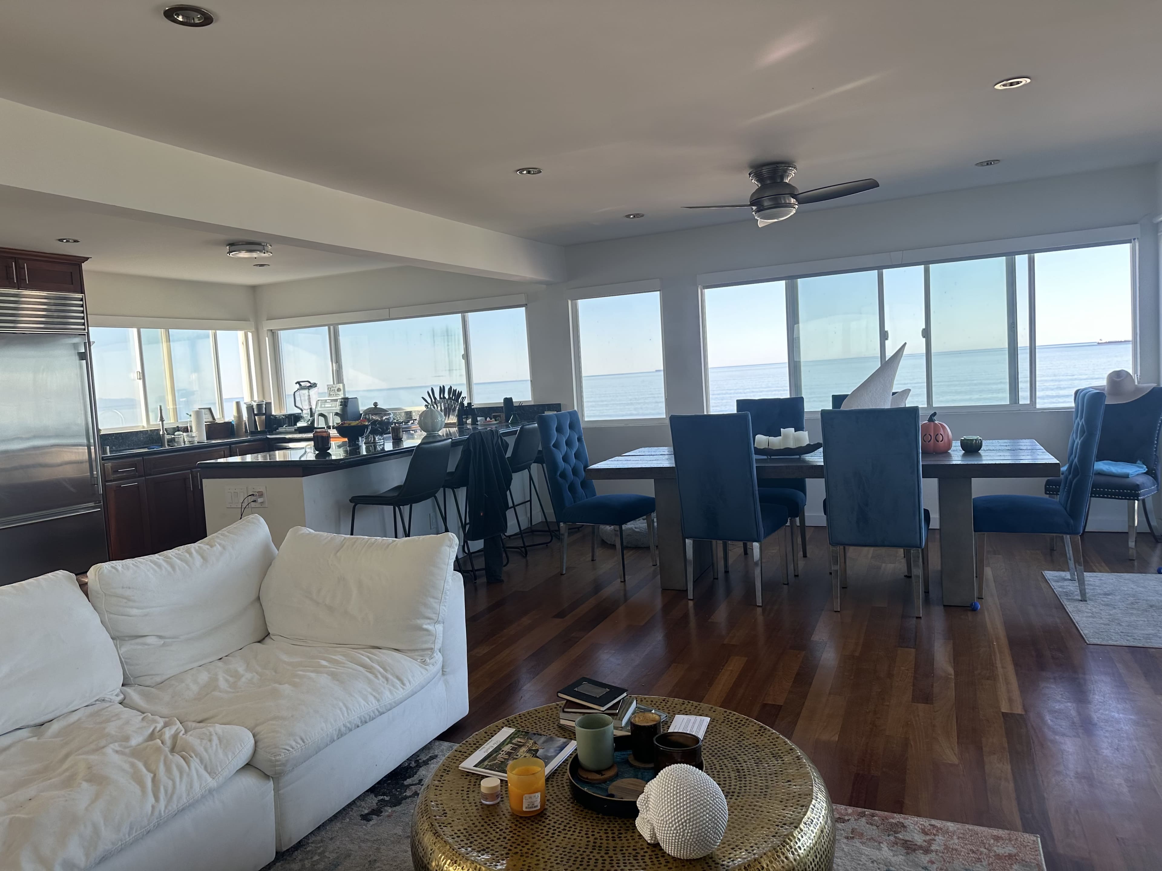 The image shows a bright, open-concept living space with a white sofa, blue dining chairs, and large windows overlooking a body of water.