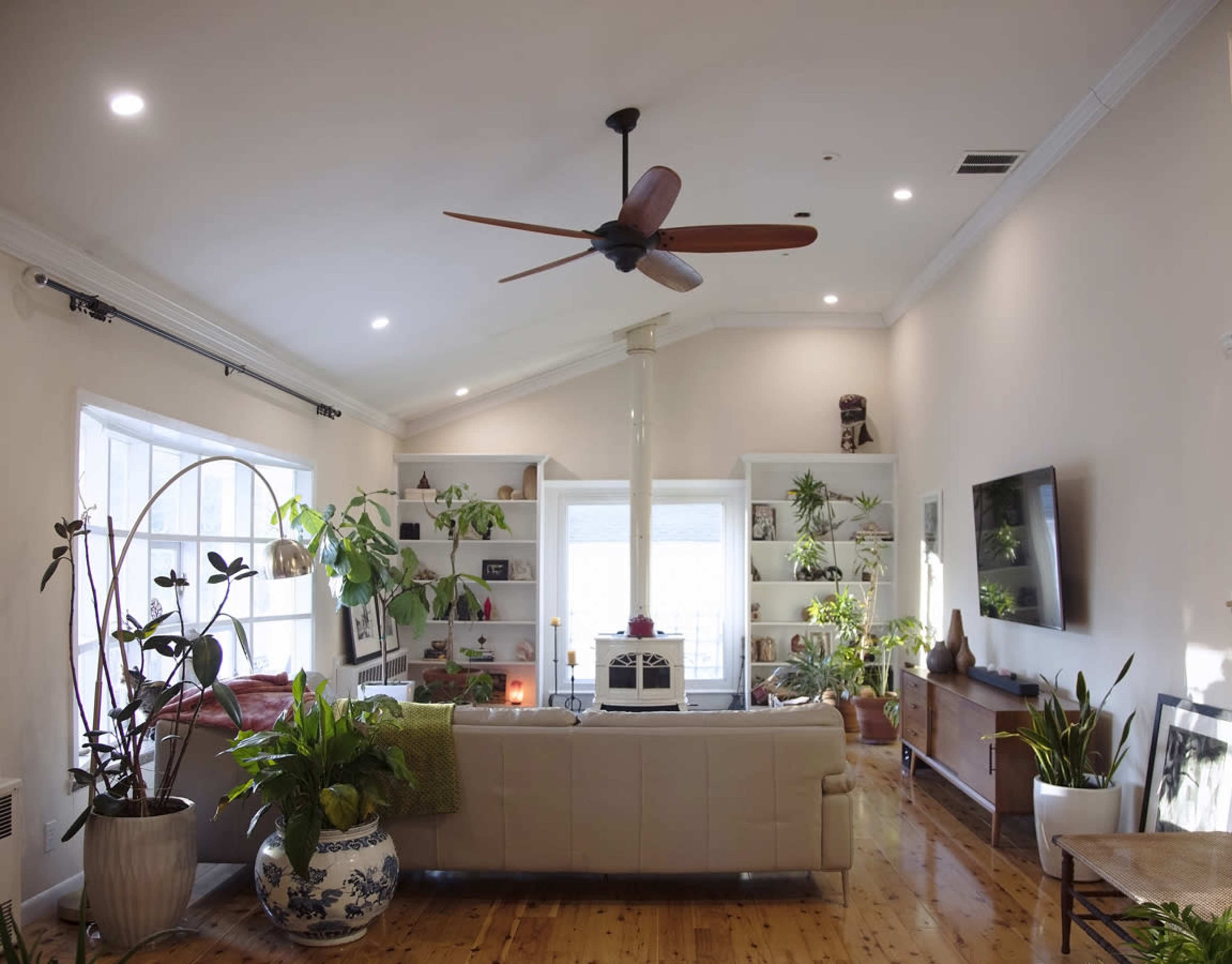 A spacious living room with a large window, a light-colored couch, houseplants, and shelves filled with decorative items.