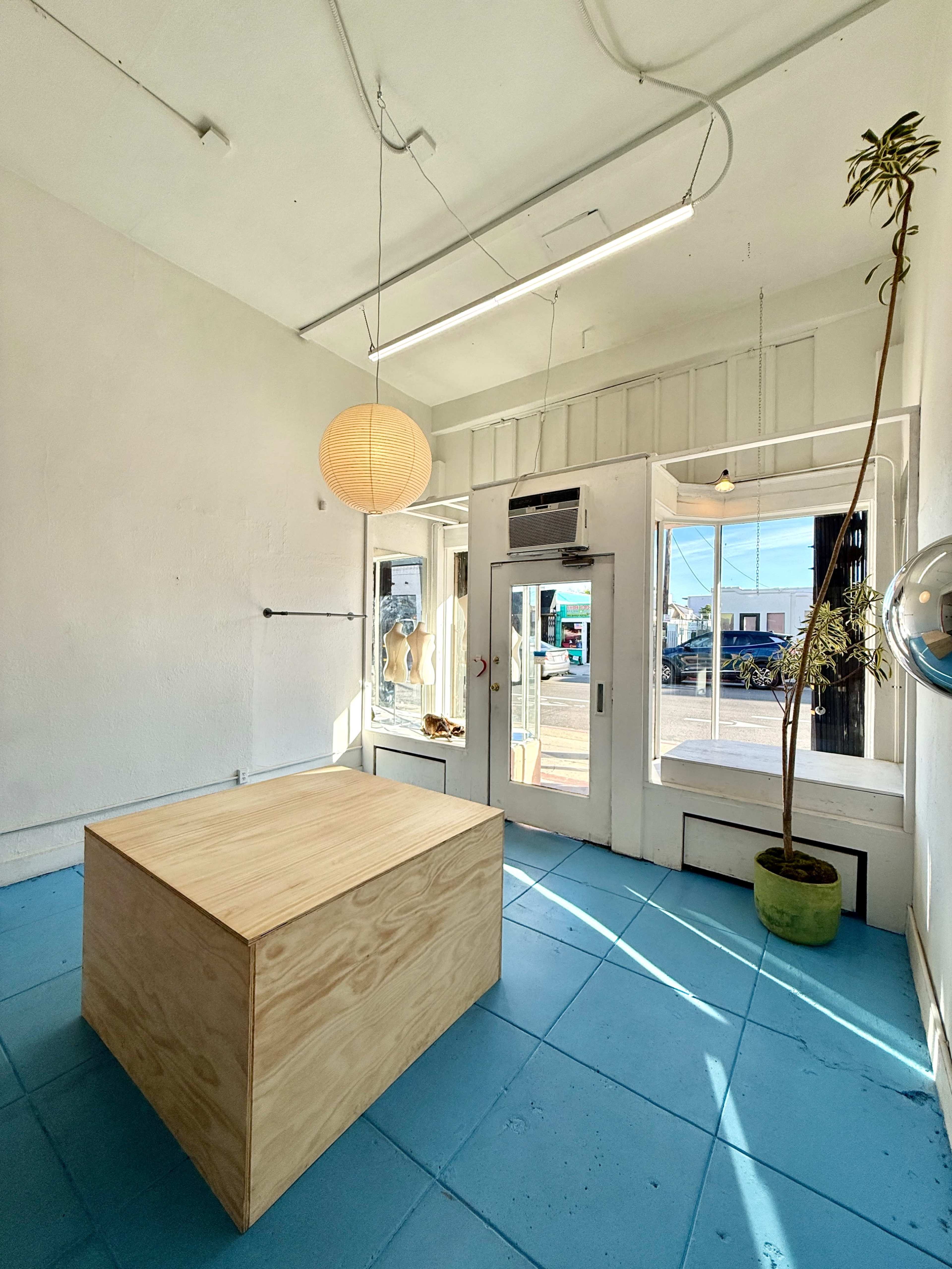 The image shows a brightly lit room with a wooden box in the center, large windows letting in natural light, and a potted plant near the entrance.