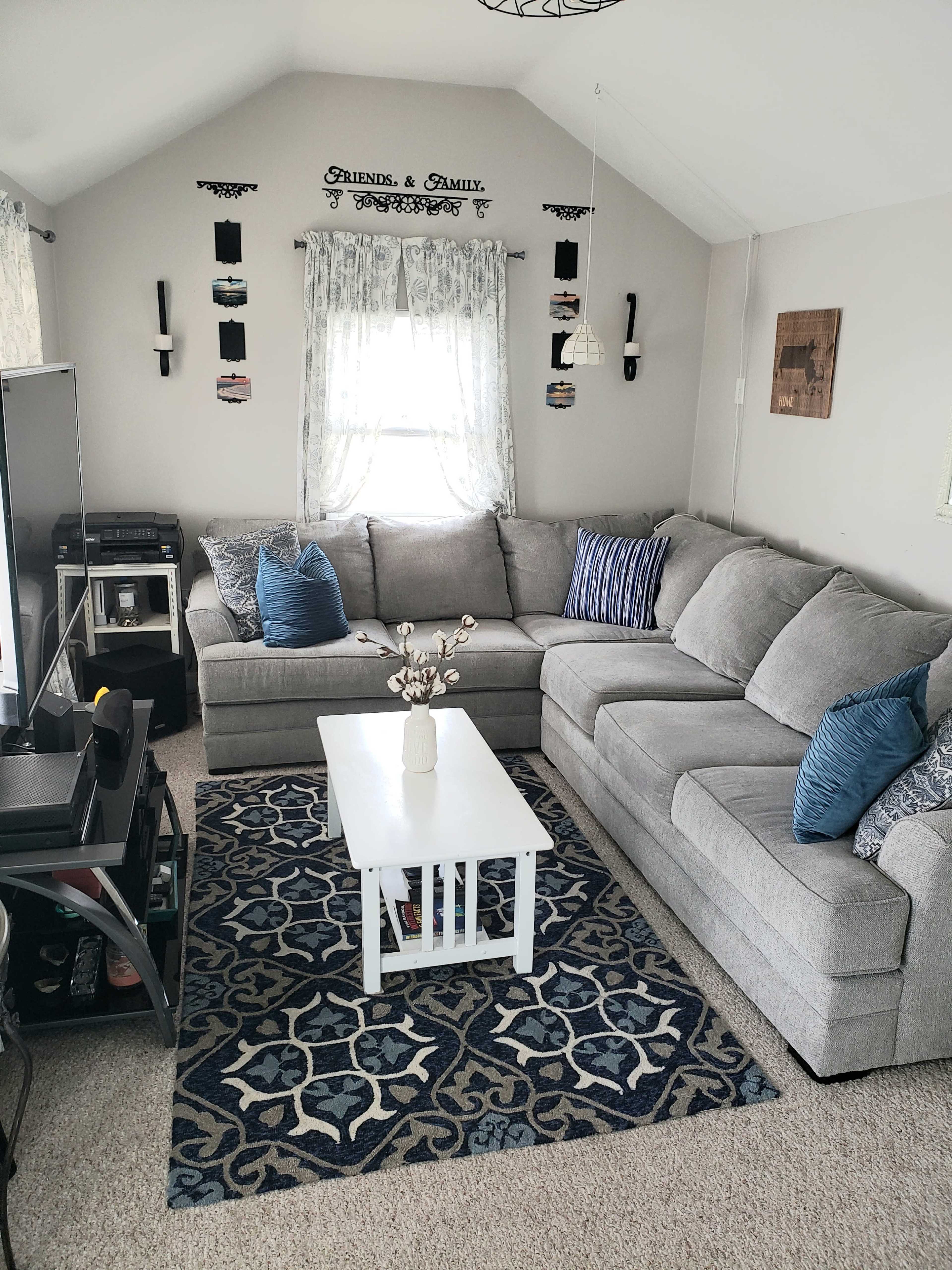 The living room features a grey sectional sofa, a white coffee table, and a patterned blue rug, with windows adorned by light curtains.
