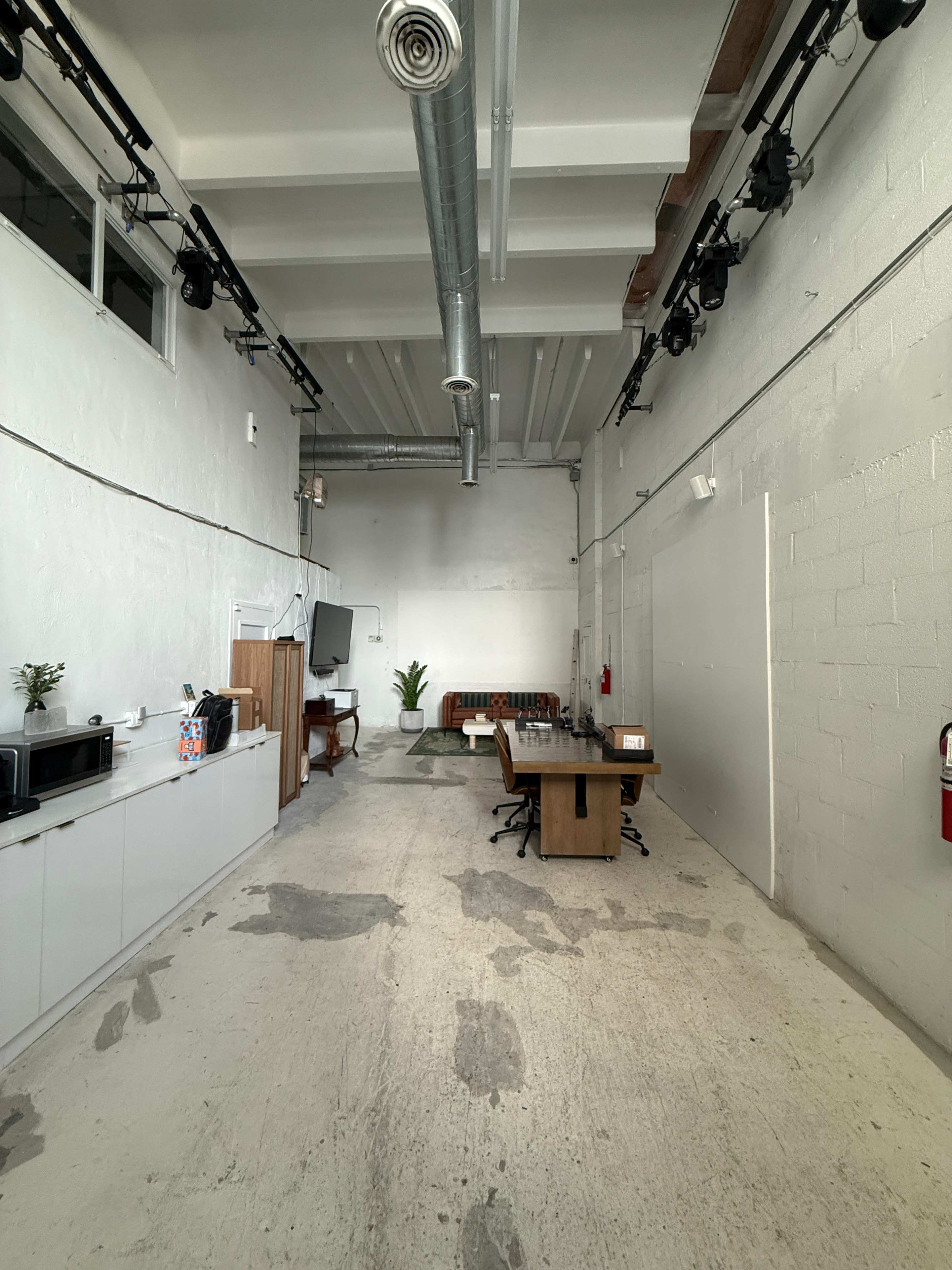 The image shows a spacious, minimalist interior of a studio or office with a long table, chairs, a couch, and a kitchenette along one wall.