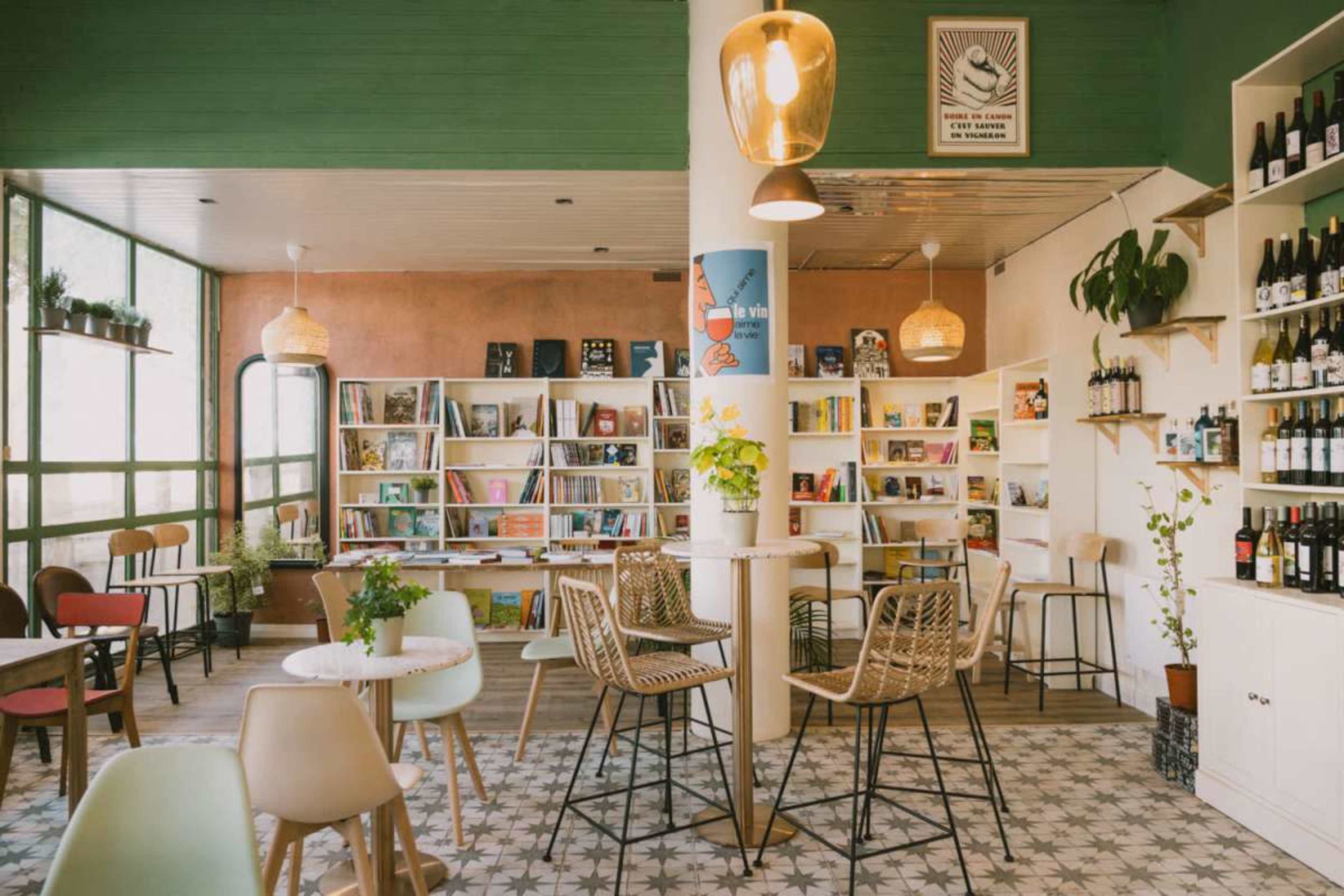 The image shows a cozy café interior featuring bookshelves, tables, and a bar area with a variety of drinks on display.