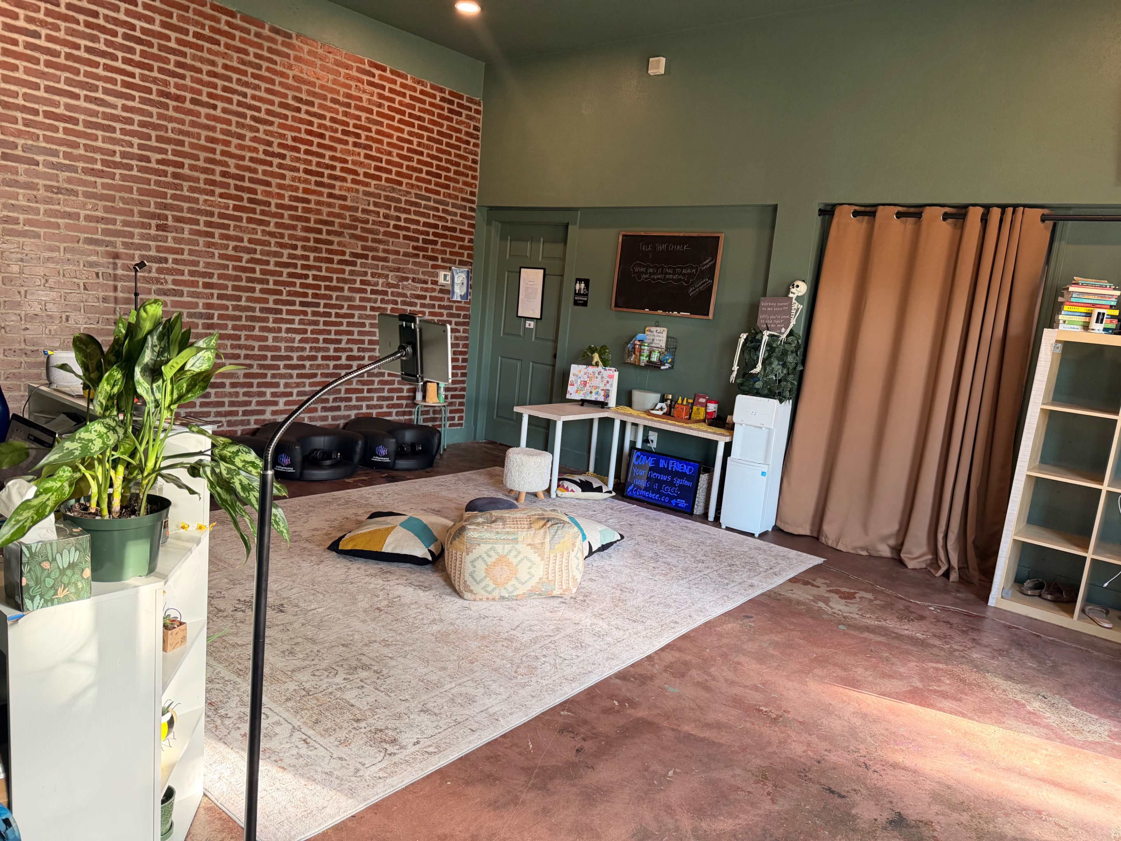 The image shows a spacious, well-lit room with a brick accent wall, a large rug, seating cushions, and shelves filled with books and decorative items.