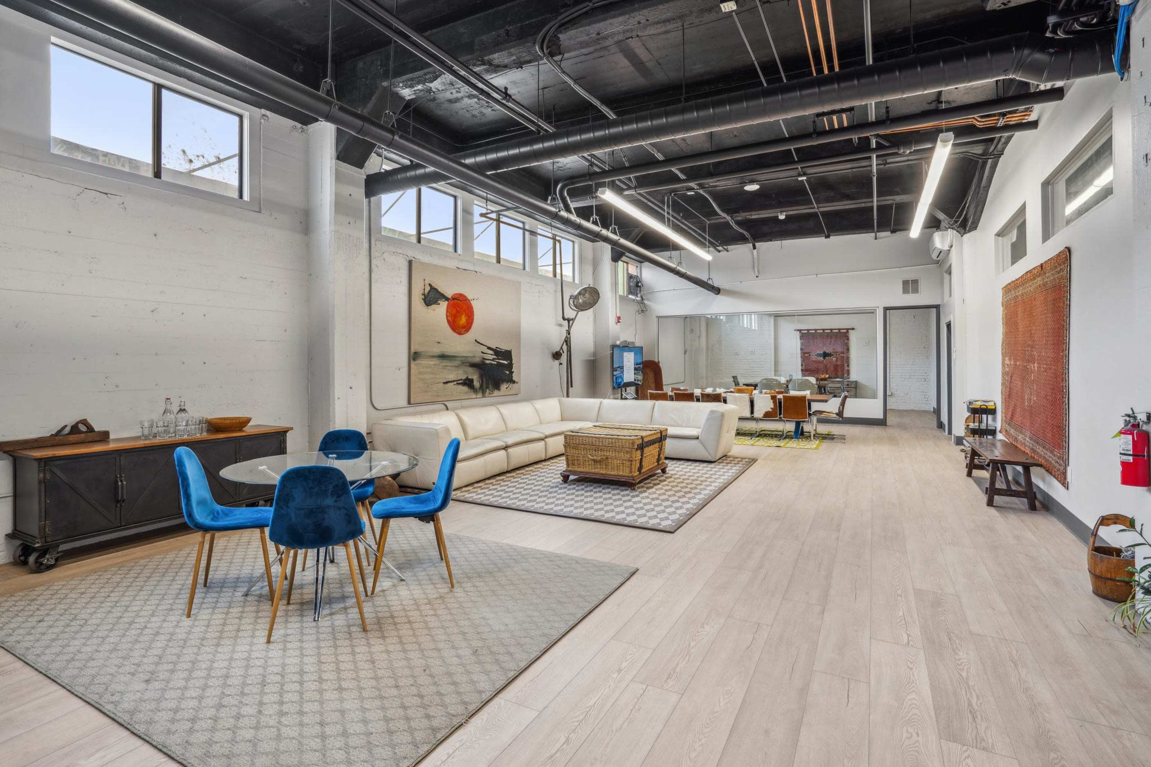 The image shows a spacious, modern living area with a dining table and chairs, a sectional sofa, and various seating arrangements along with artwork on the walls.