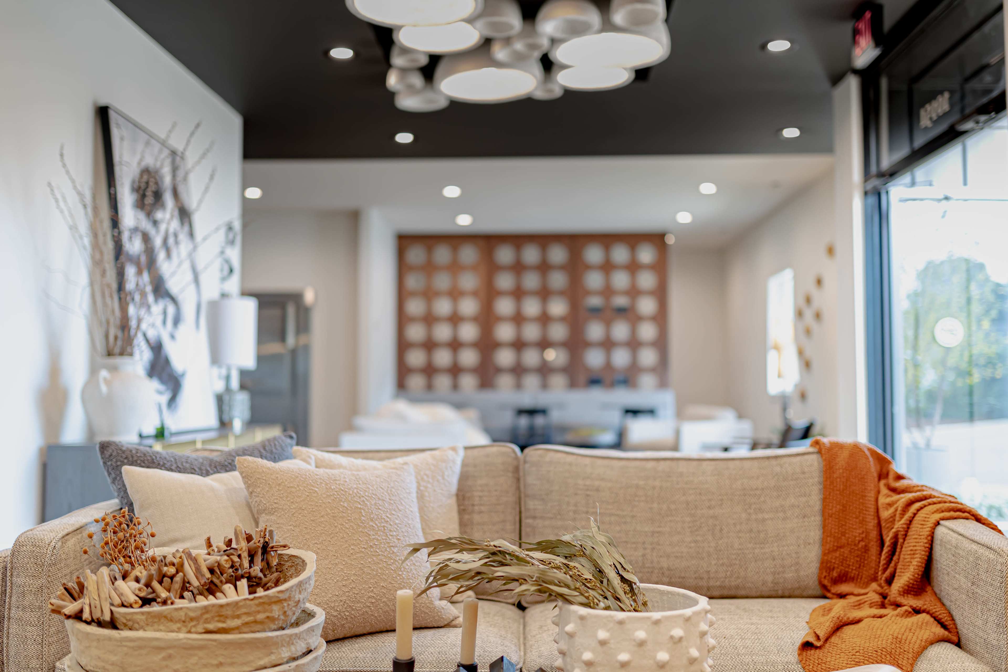 A comfortable living space features a beige couch adorned with pillows, a decorative bowl of twigs, candles, and an orange throw, with a bright, modern room visible in the background.