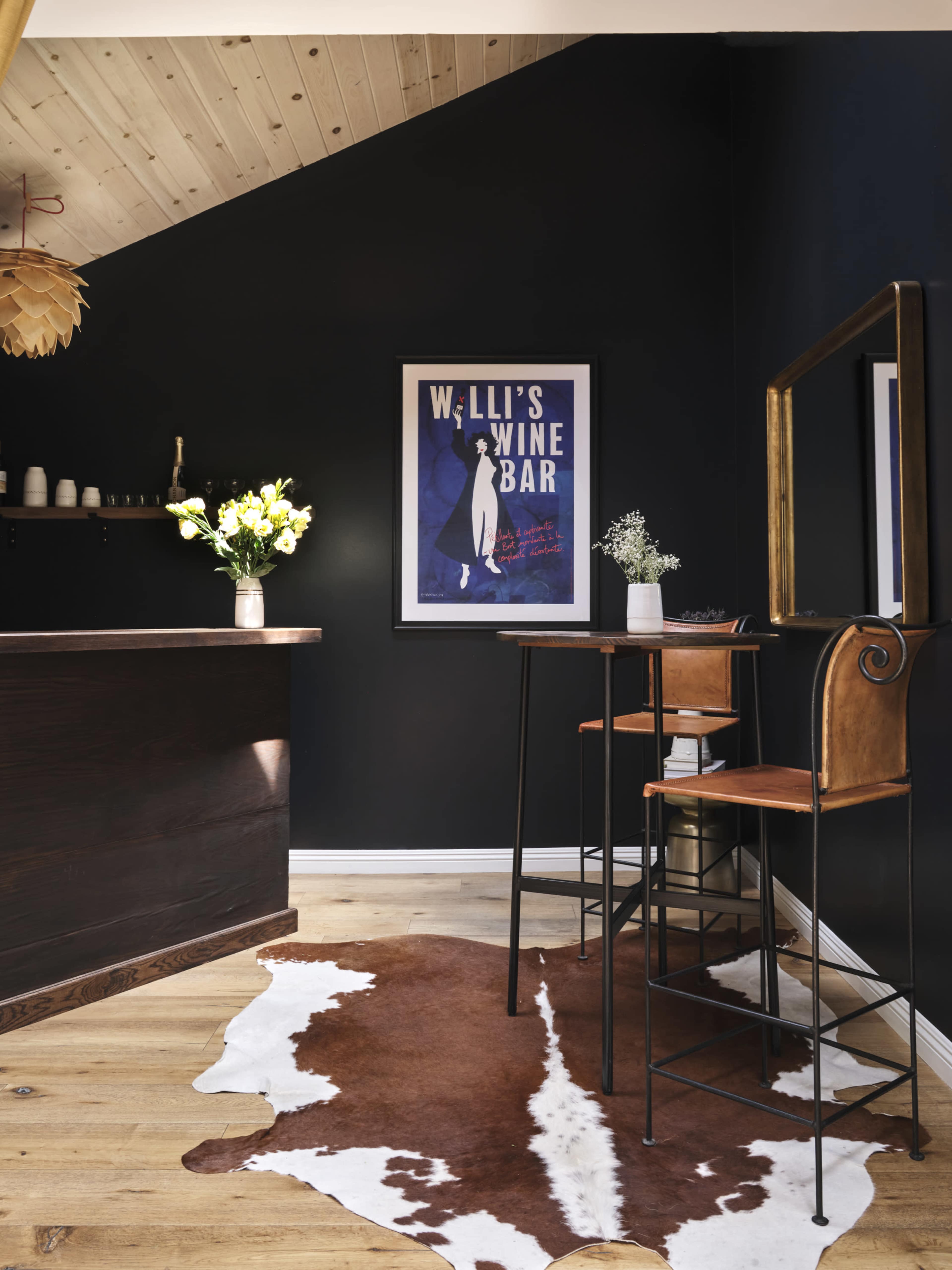 A modern bar area with a dark-painted wall, a framed poster, a wooden counter, and a tall table with metal and leather stools on a cowhide rug.