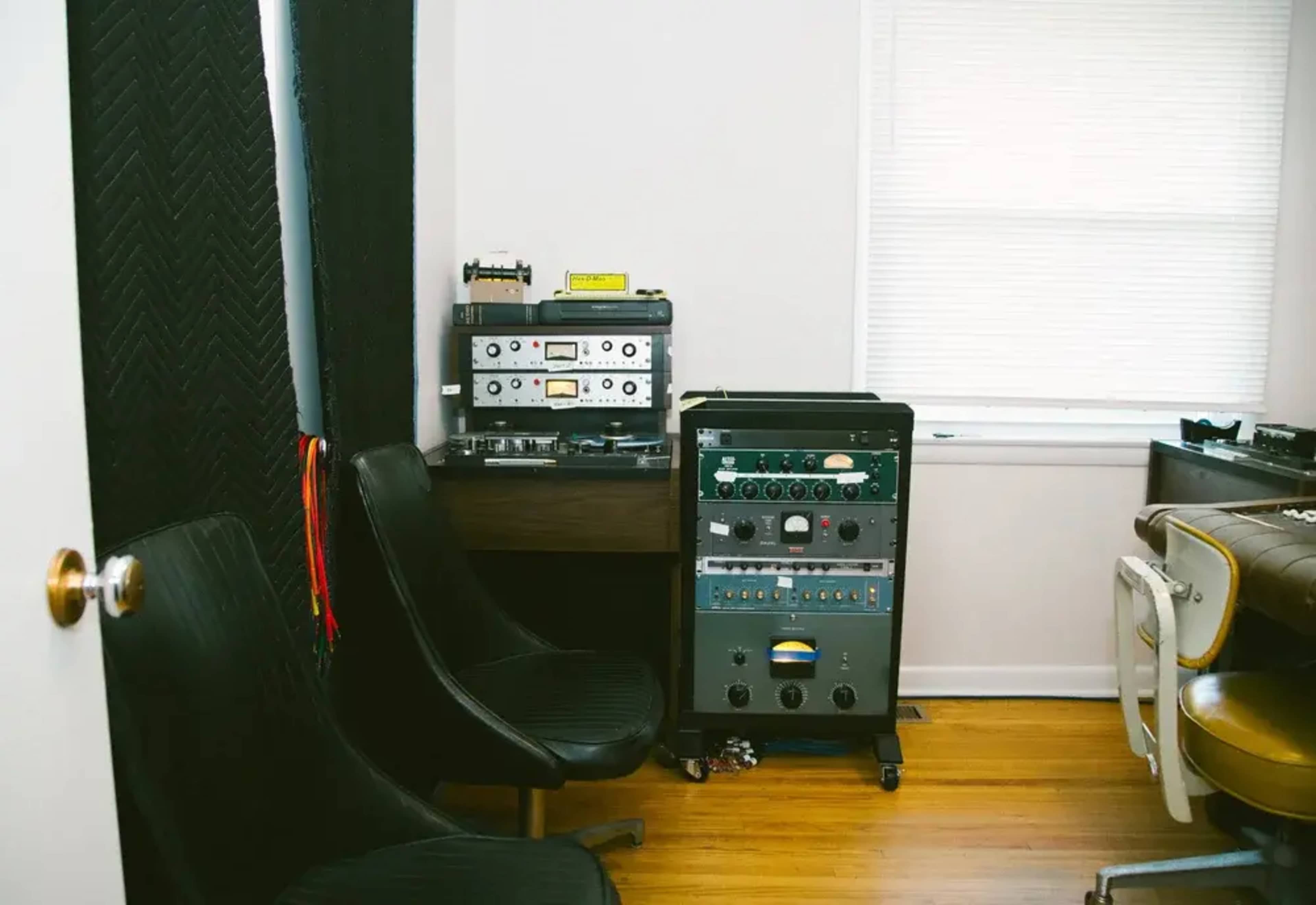The image shows a room with two black chairs, a window with blinds, and audio equipment including a tape machine and a rack-mounted unit on a wheeled stand.