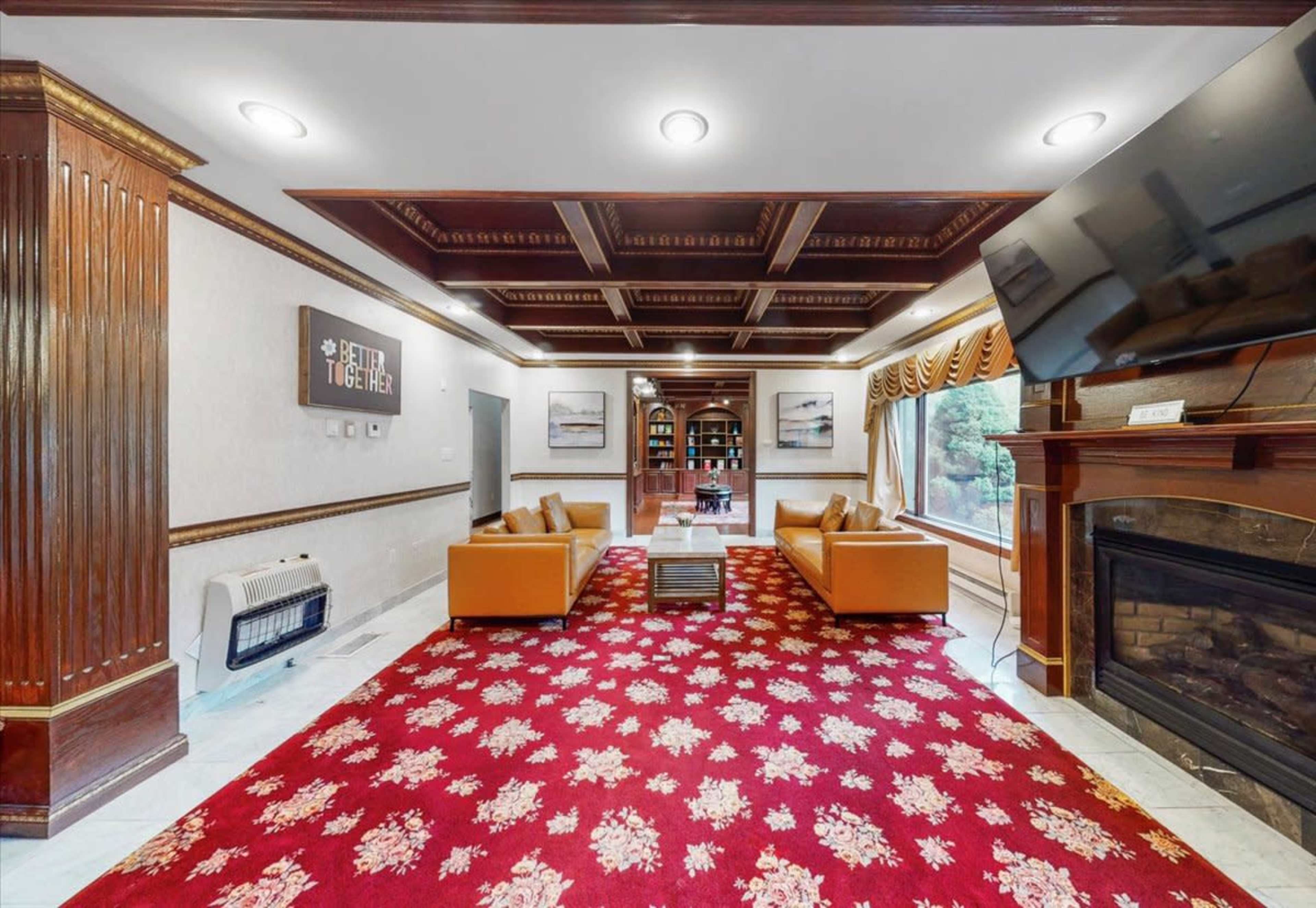 The image shows a cozy living room with orange sofas arranged around a coffee table, a large TV mounted on the wall, and a decorative red carpet.