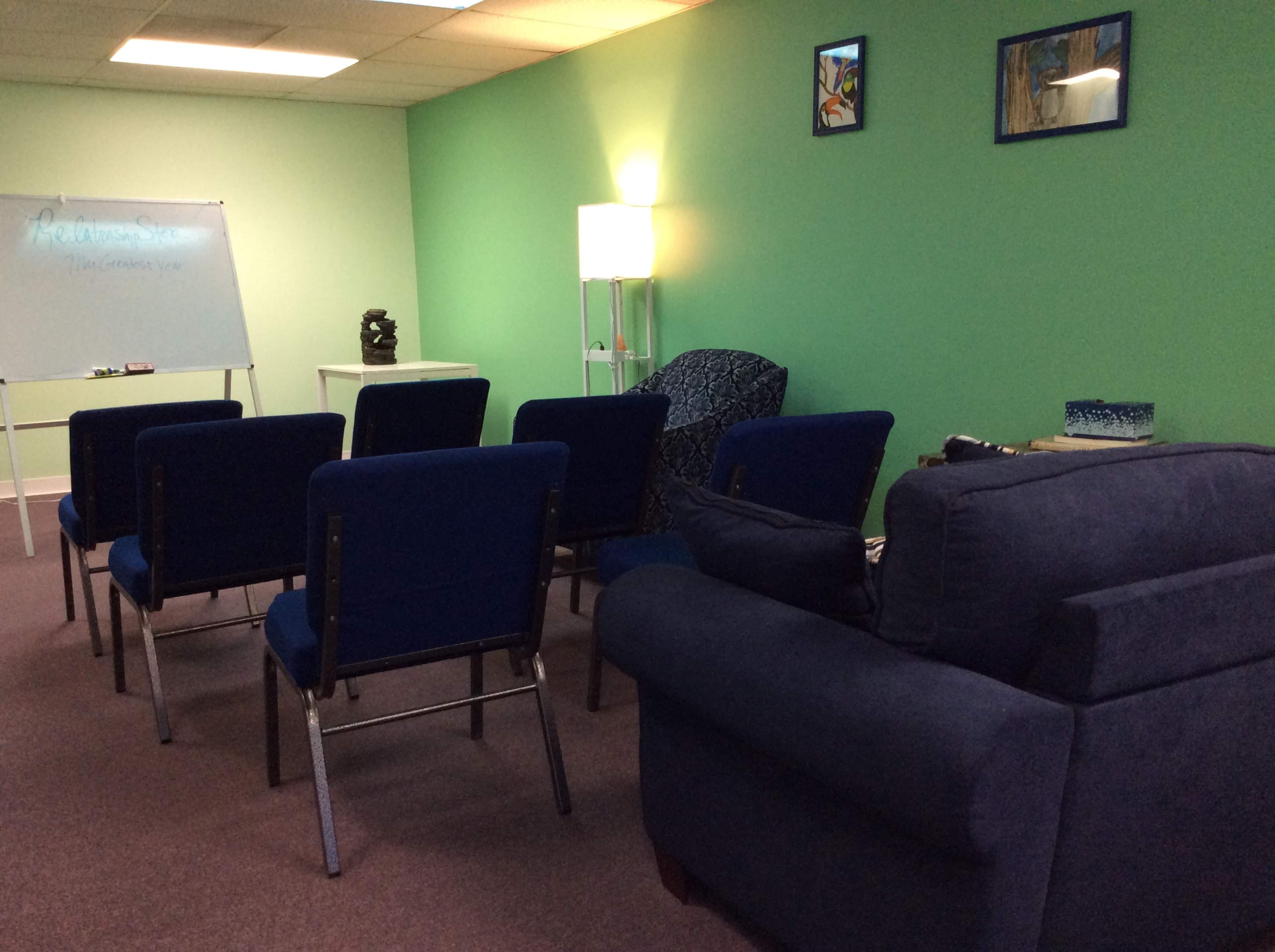 A small meeting room with blue chairs arranged in a semicircle facing a whiteboard, alongside a couch and a decorative lamp against a green wall.