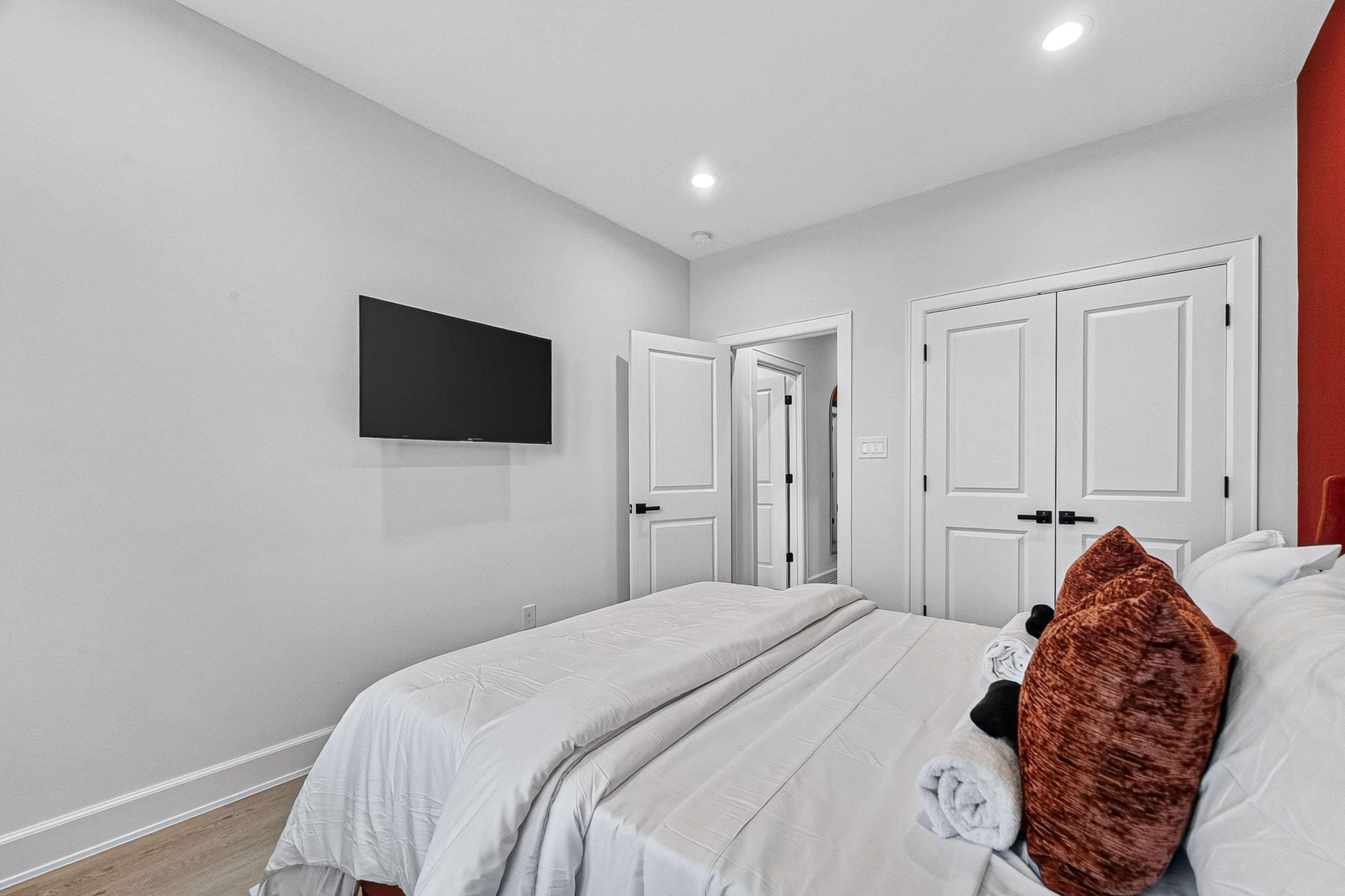 A neatly arranged bedroom features a bed with white linens, a flat-screen TV mounted on the wall, and double doors leading to another room.