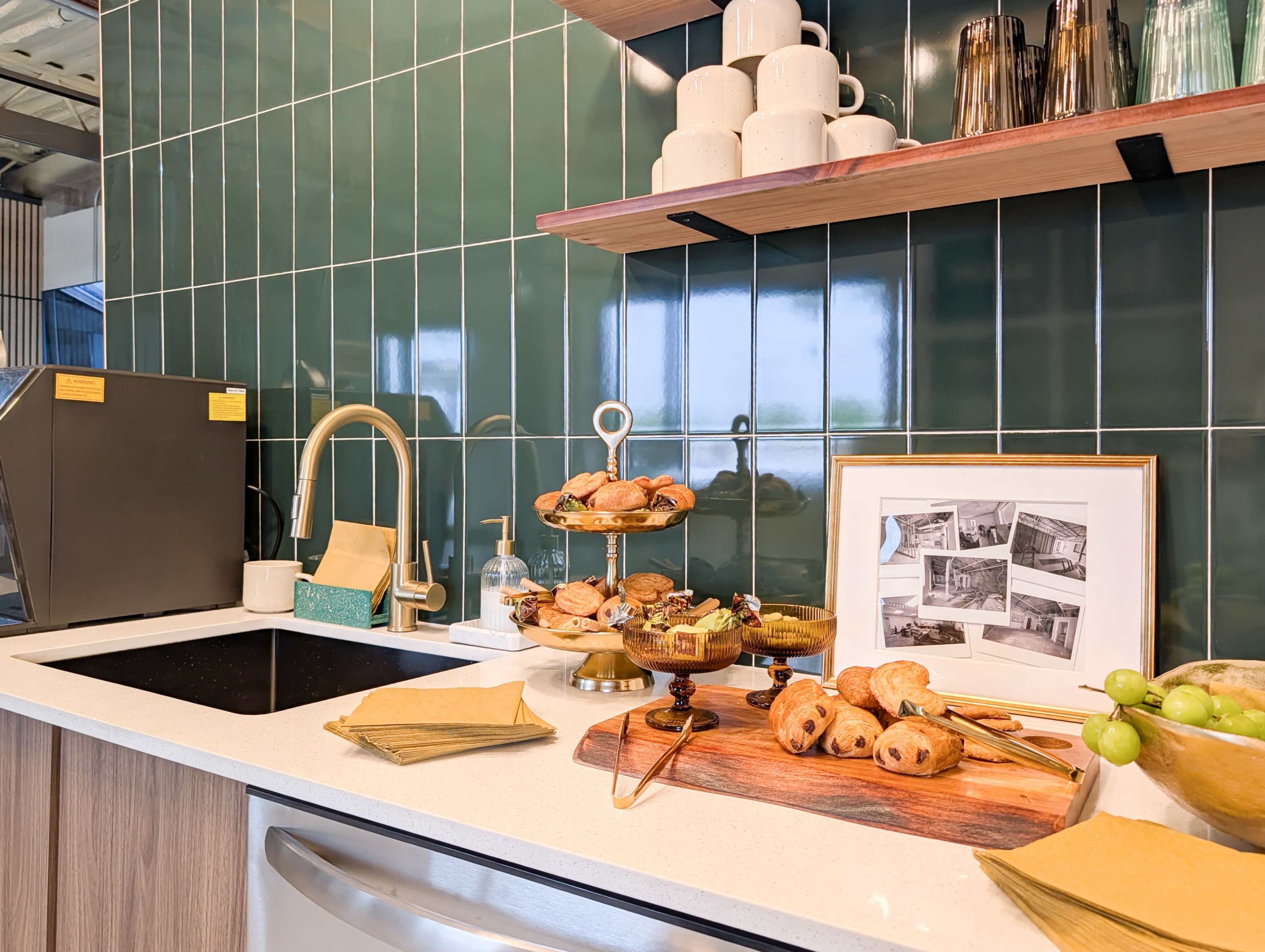 The image shows a modern kitchen countertop with an array of baked goods displayed on a tiered stand, along with a framed set of black-and-white photos and kitchen utensils.