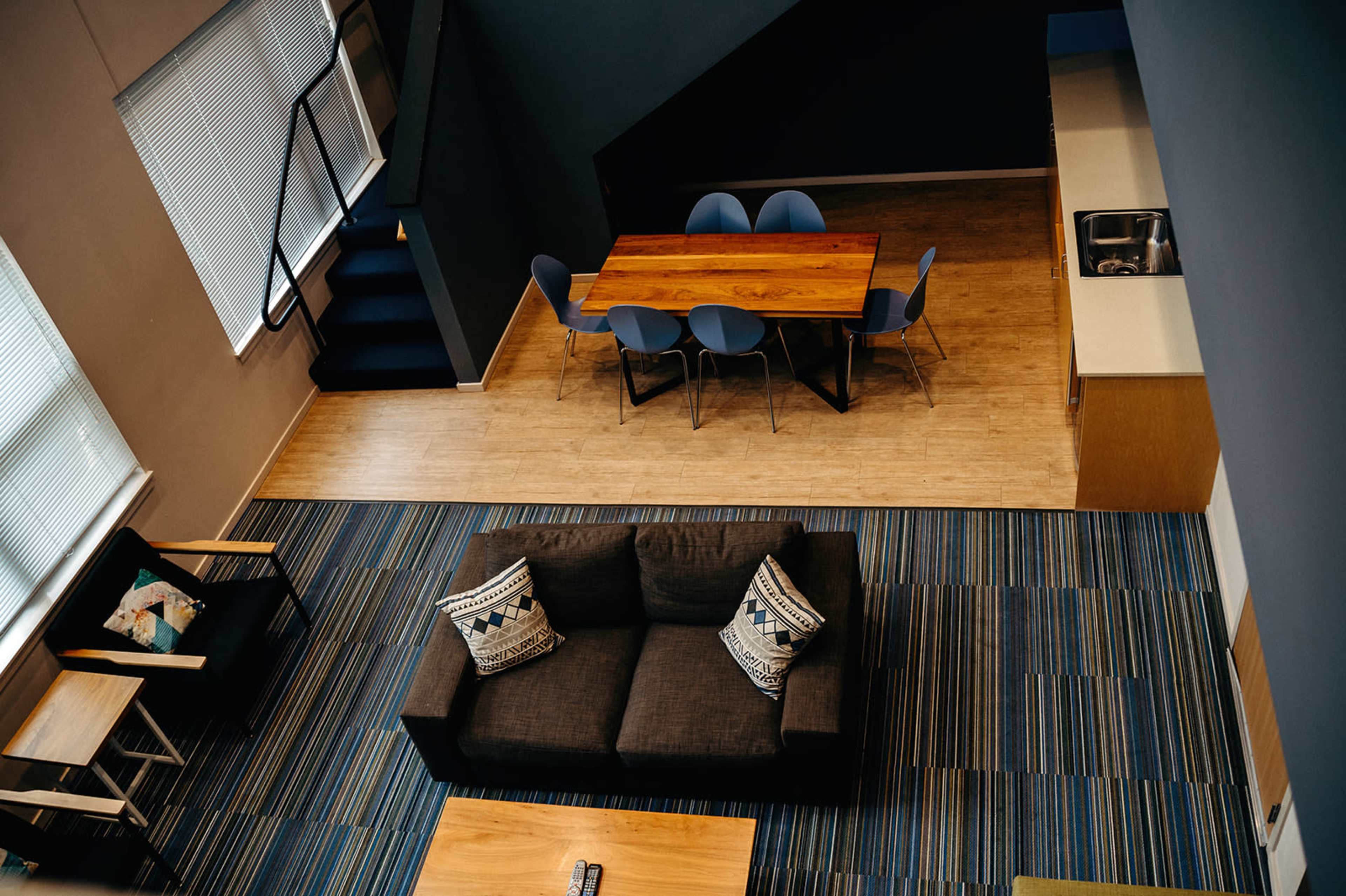 A modern living space featuring a dark sofa with decorative pillows, a wooden dining table surrounded by blue chairs, and a kitchen area, all set against a backdrop of striped flooring.