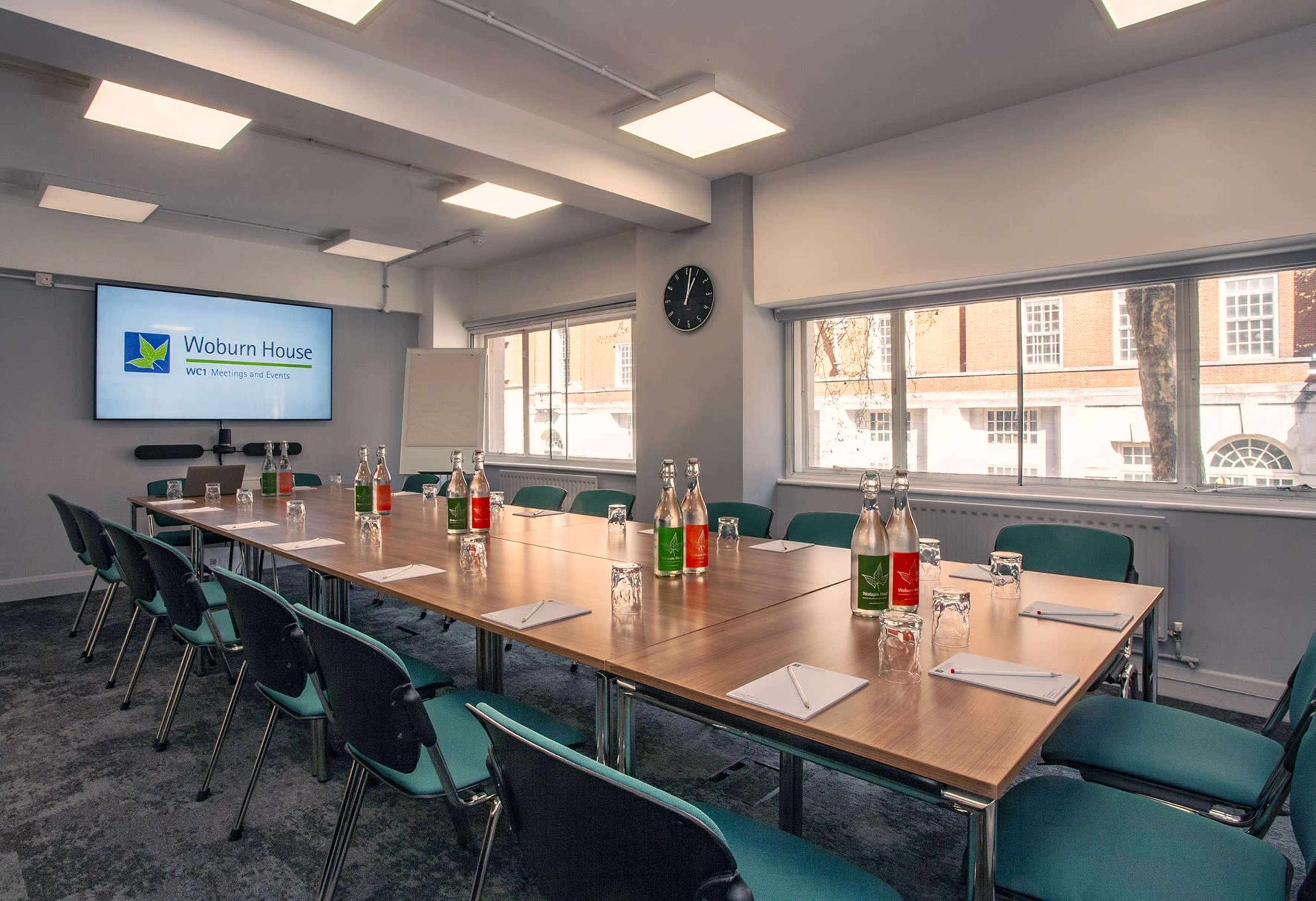A meeting room with a large rectangular table set for a conference, featuring water bottles, notepads, and a presentation screen at one end.