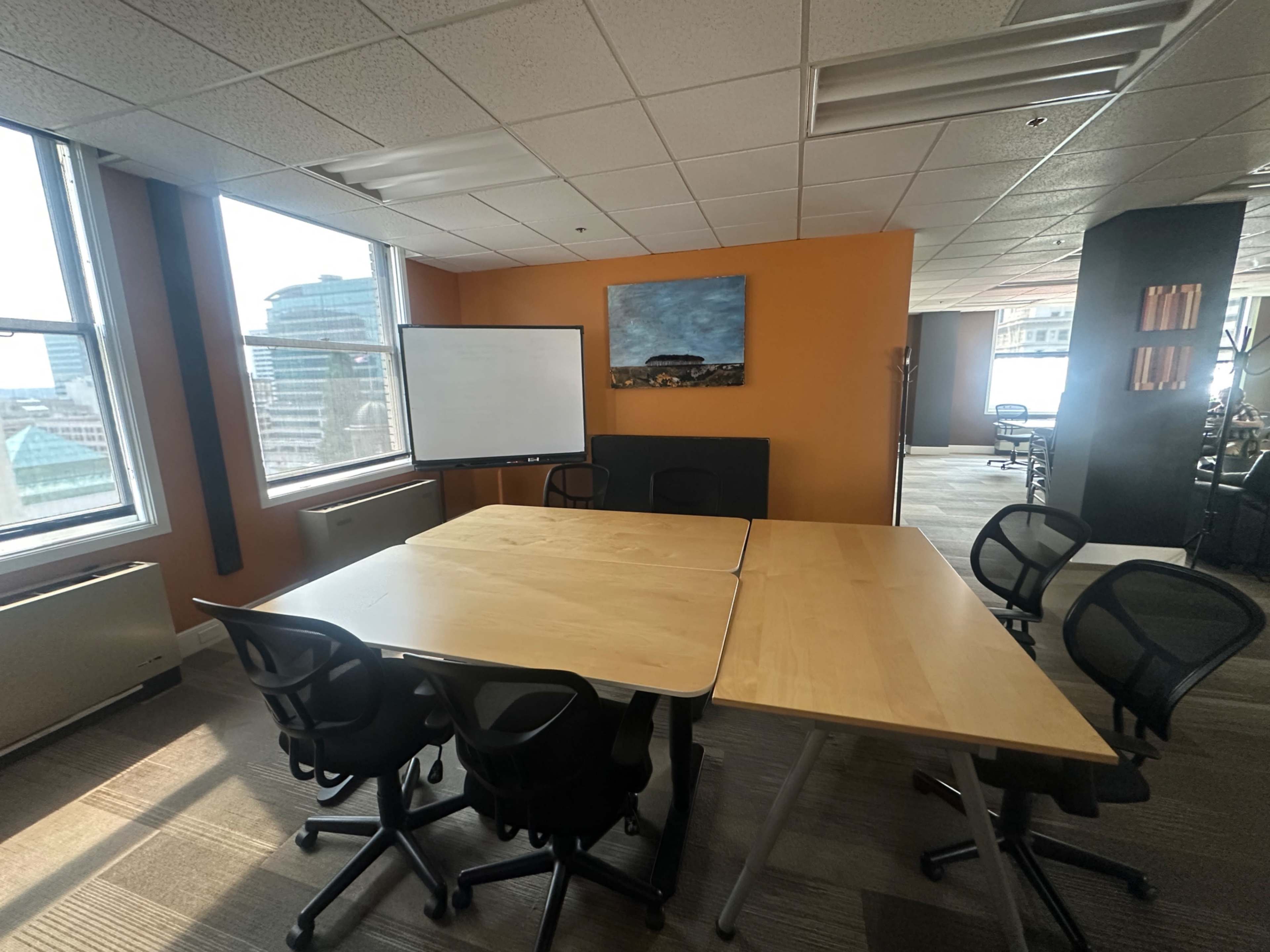The image shows a modern conference room with a large wooden table, several black office chairs, a whiteboard, and large windows allowing natural light to enter the space.