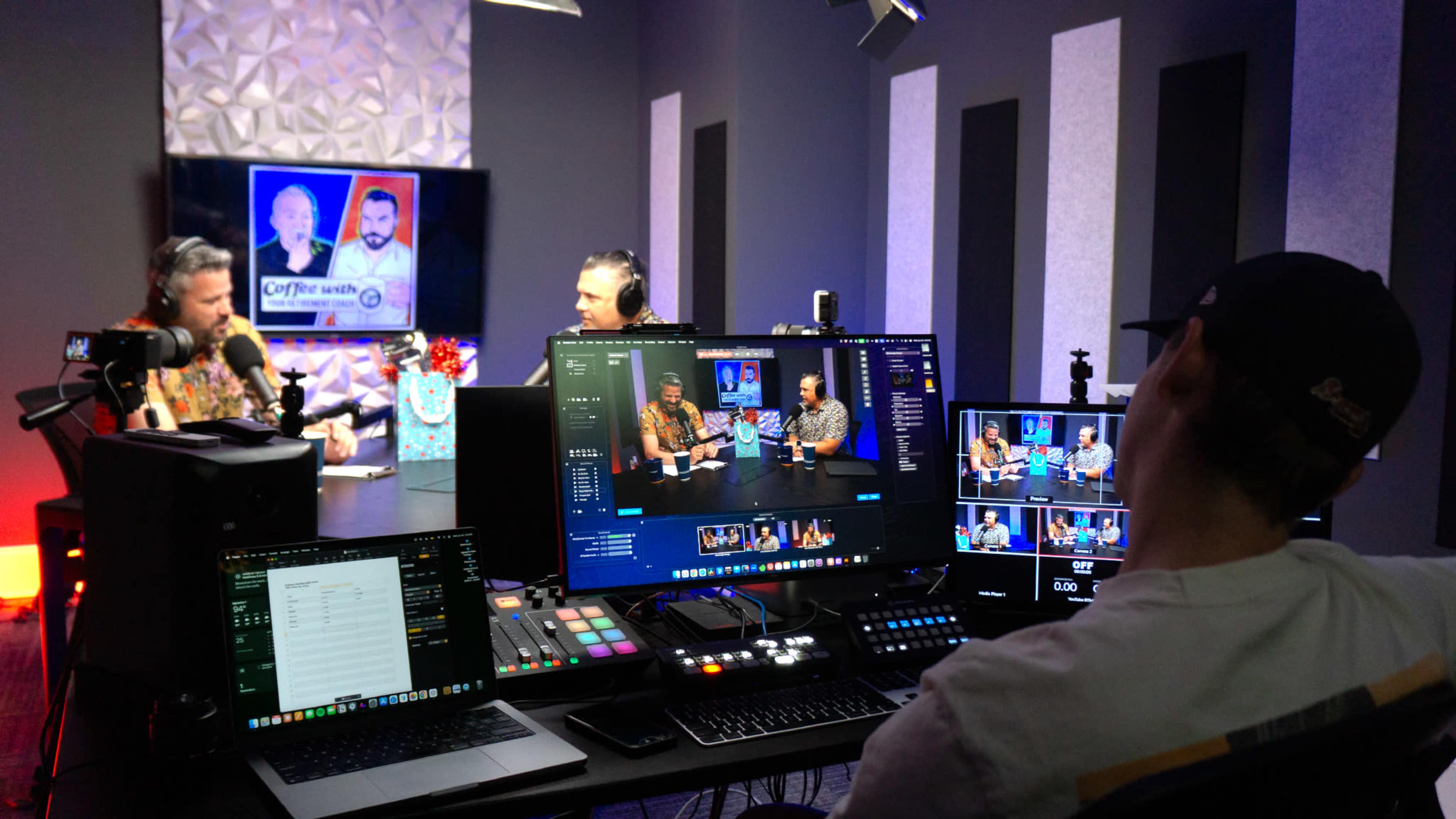 The image shows a video recording studio with two hosts seated at a table discussing, while a technical operator manages equipment and monitors multiple screens.