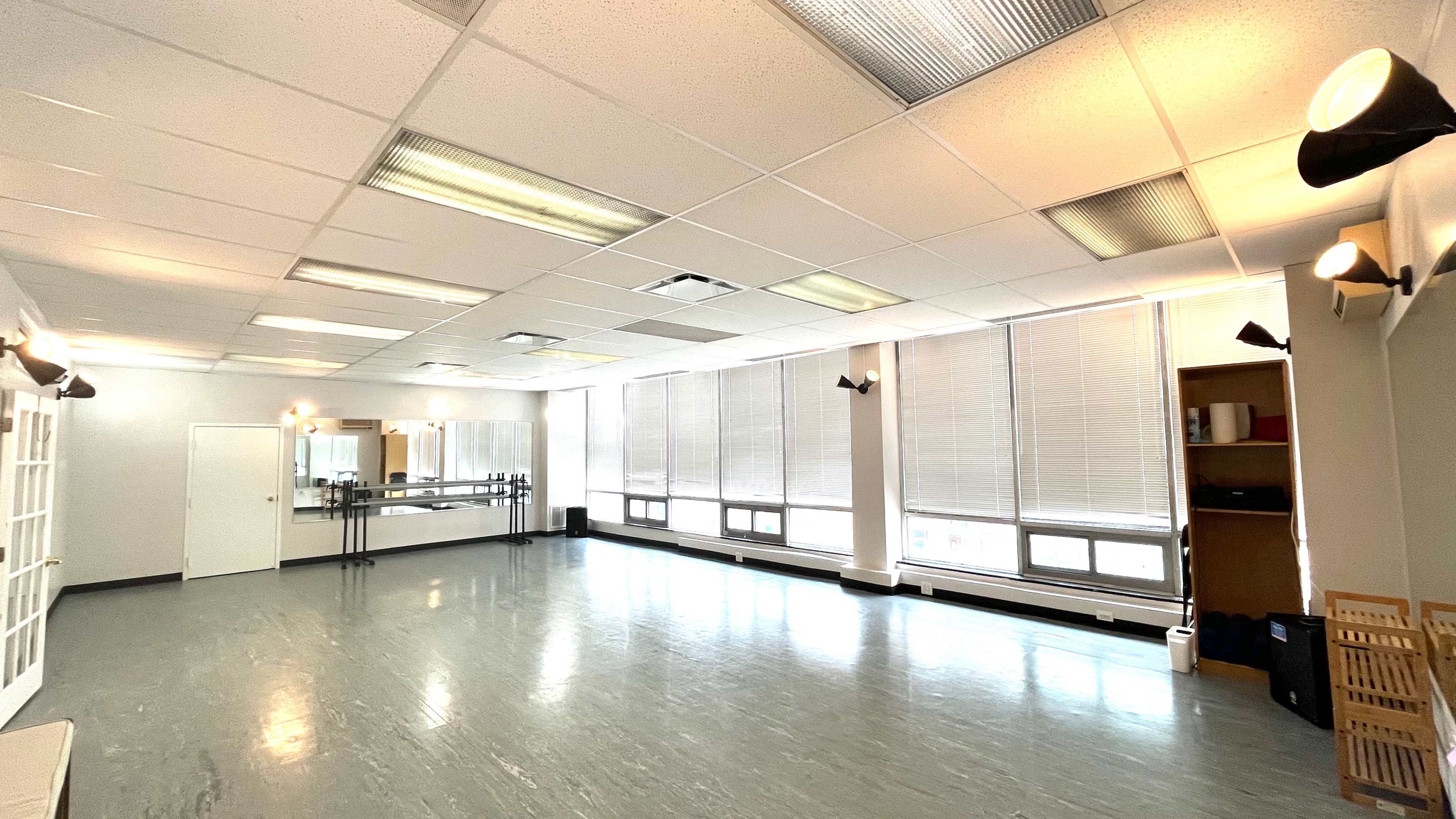A spacious dance studio with large windows, grey flooring, mirrors on one wall, and adjustable lighting fixtures.