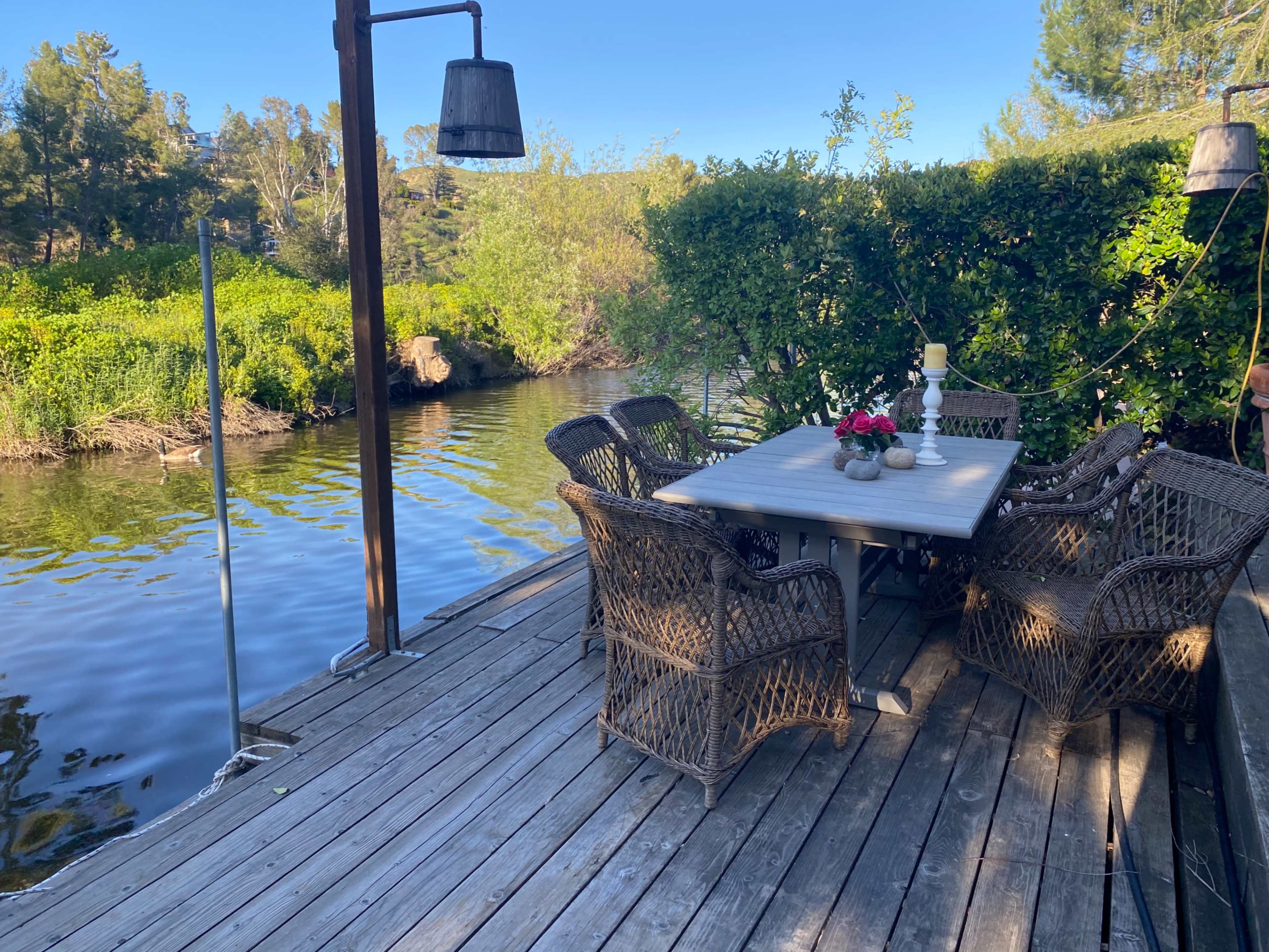 A wooden deck features a table surrounded by chairs, overlooking a serene riverbank with lush greenery.