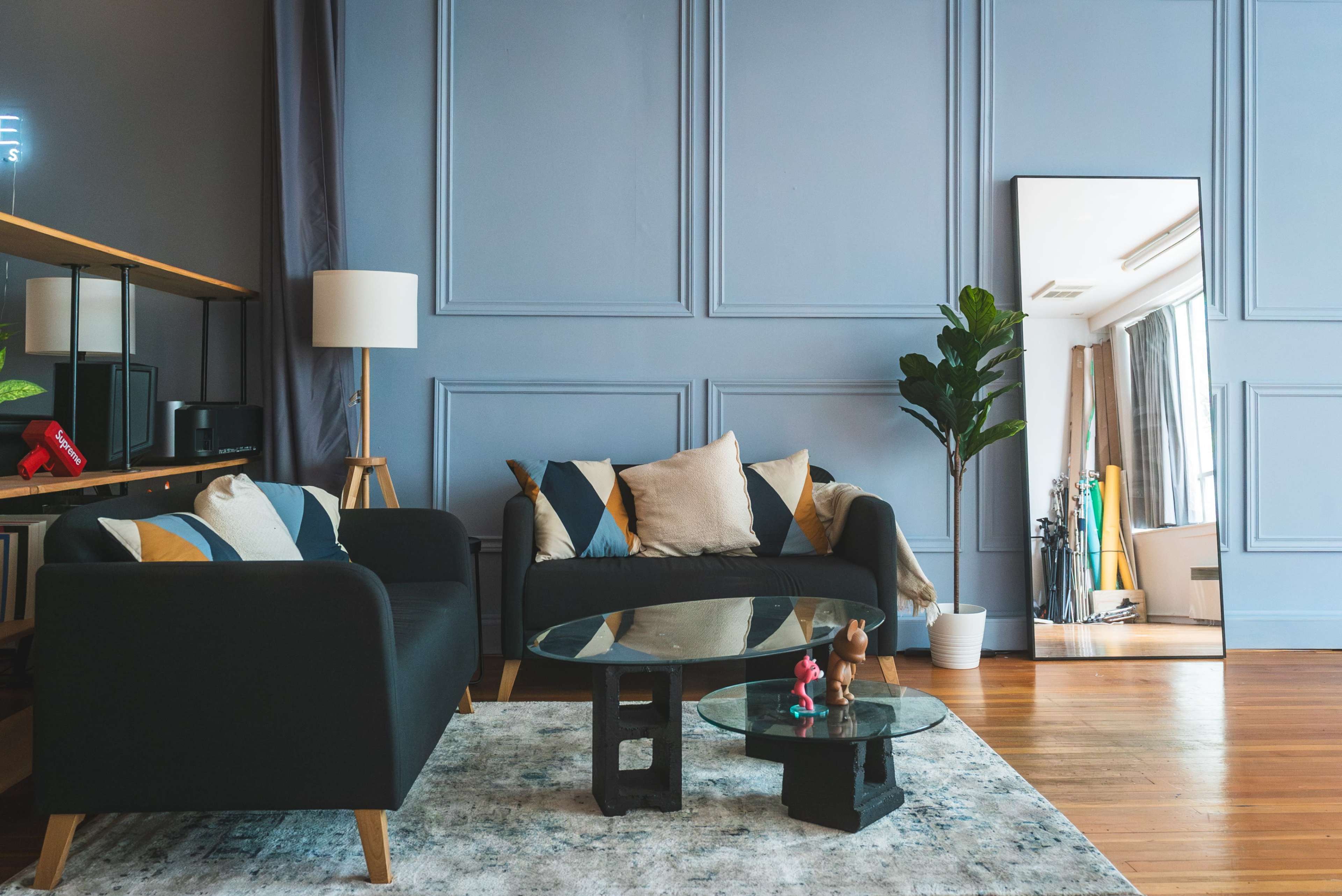 A living room featuring a black couch with decorative pillows, a glass coffee table, a large mirror, and a potted plant against a blue wall.