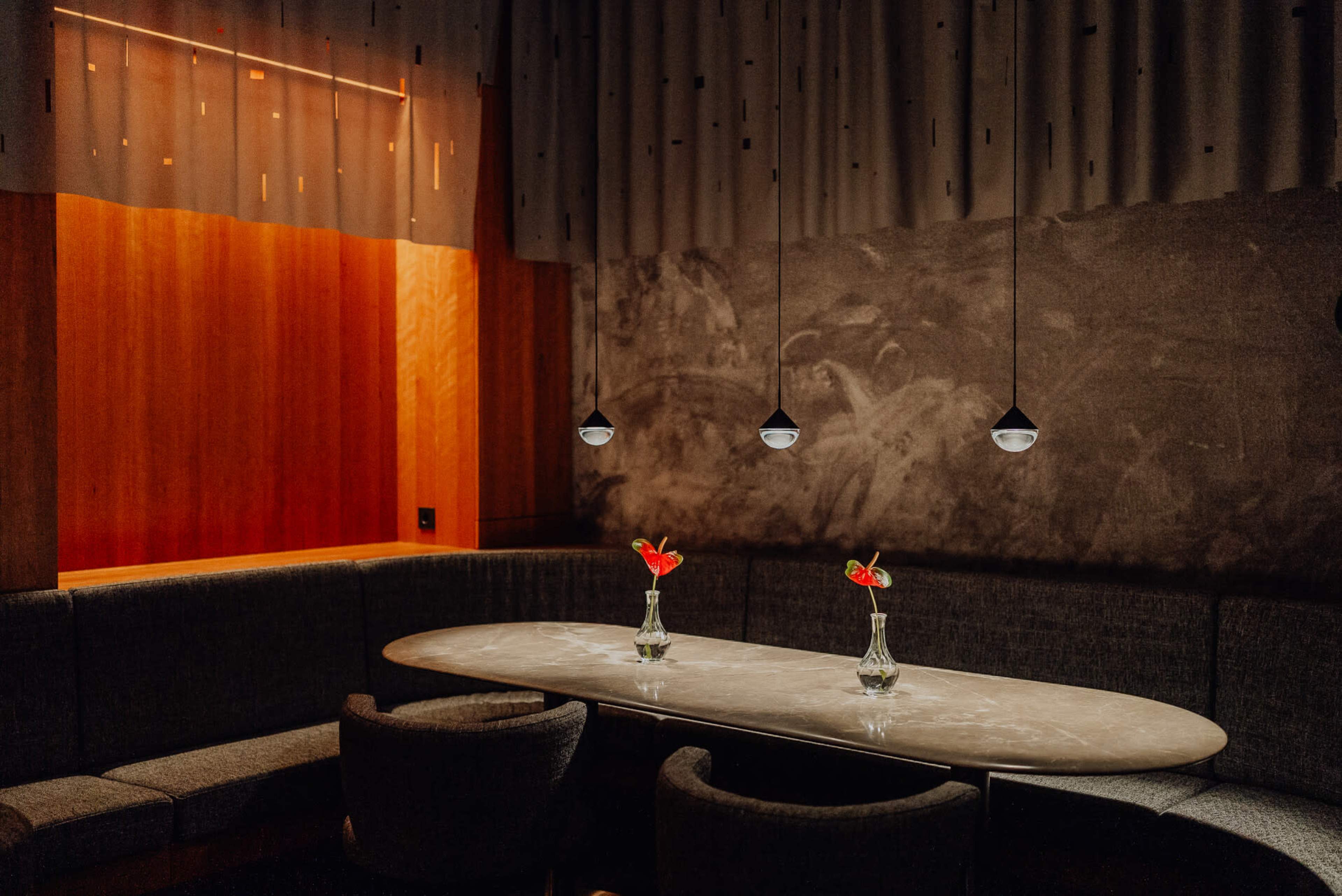 A modern lounge area features an oval marble table with two vases holding flowers, illuminated by pendant lights in a softly lit setting.